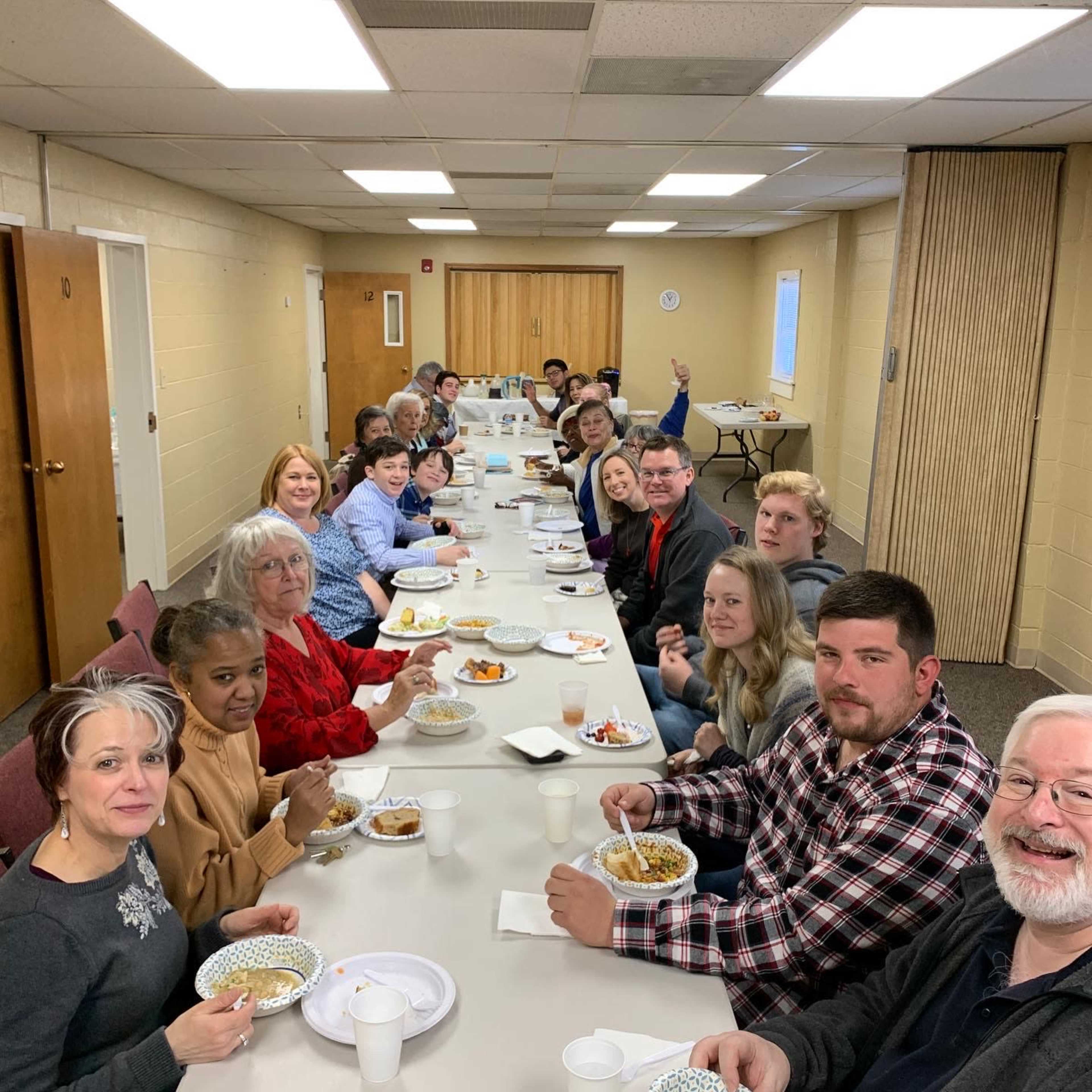 A group of people gathers around a long table, enjoying a meal together in a well-lit room.