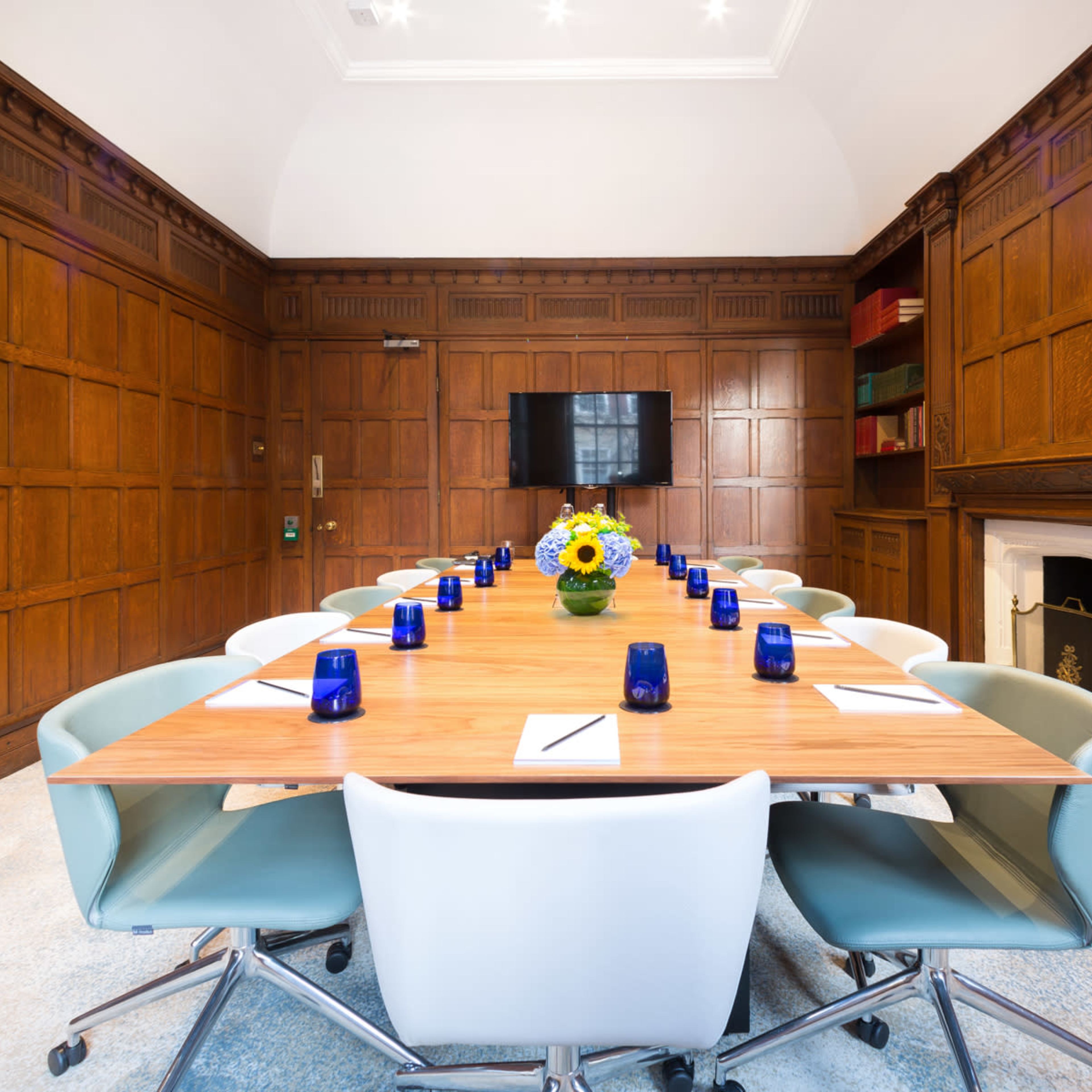 A long wooden conference table is set with blue candles and notepads in a room featuring dark wooden paneling and a large screen on the wall.