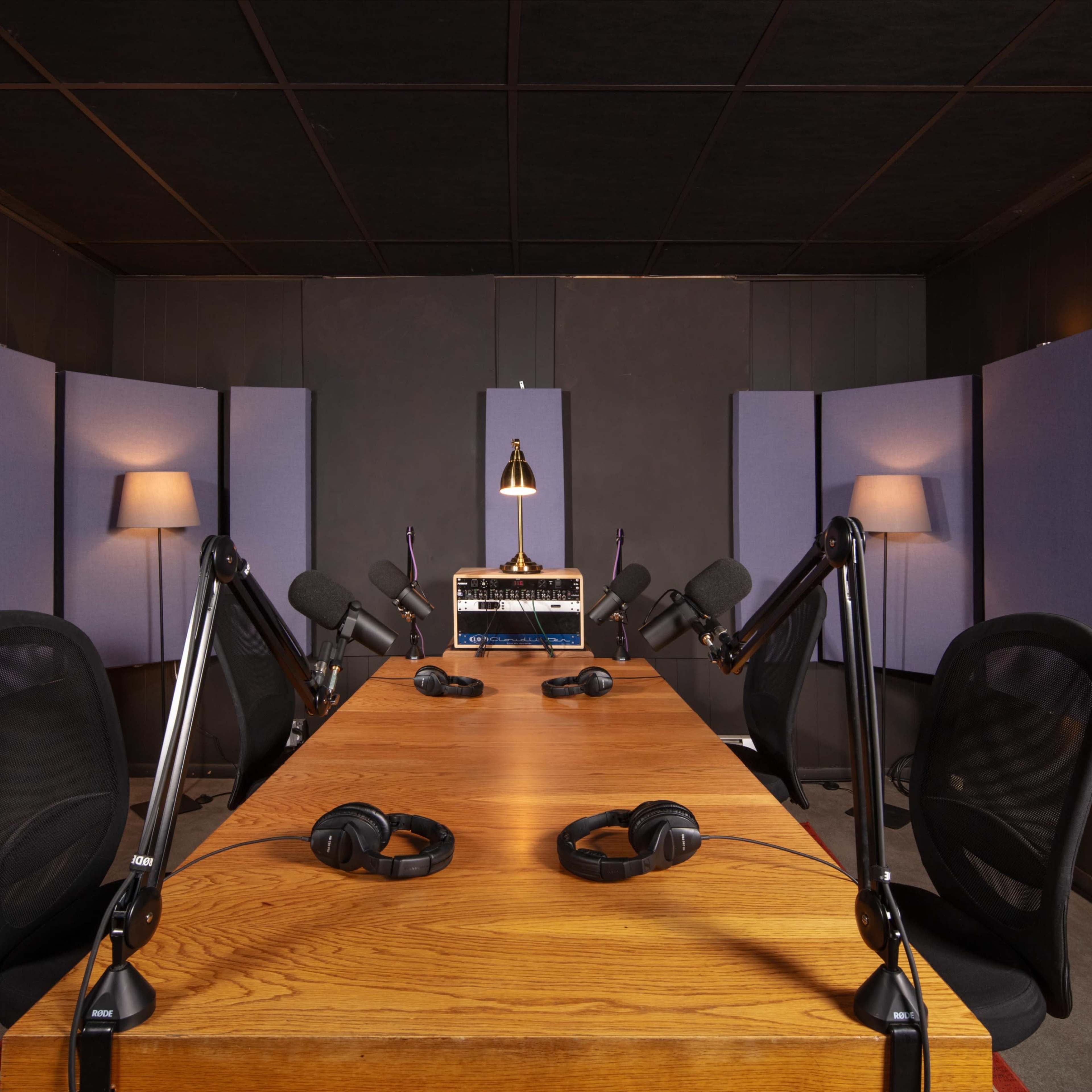 A recording studio features a large wooden table surrounded by equipped microphones, headphones, and soundproof wall panels.