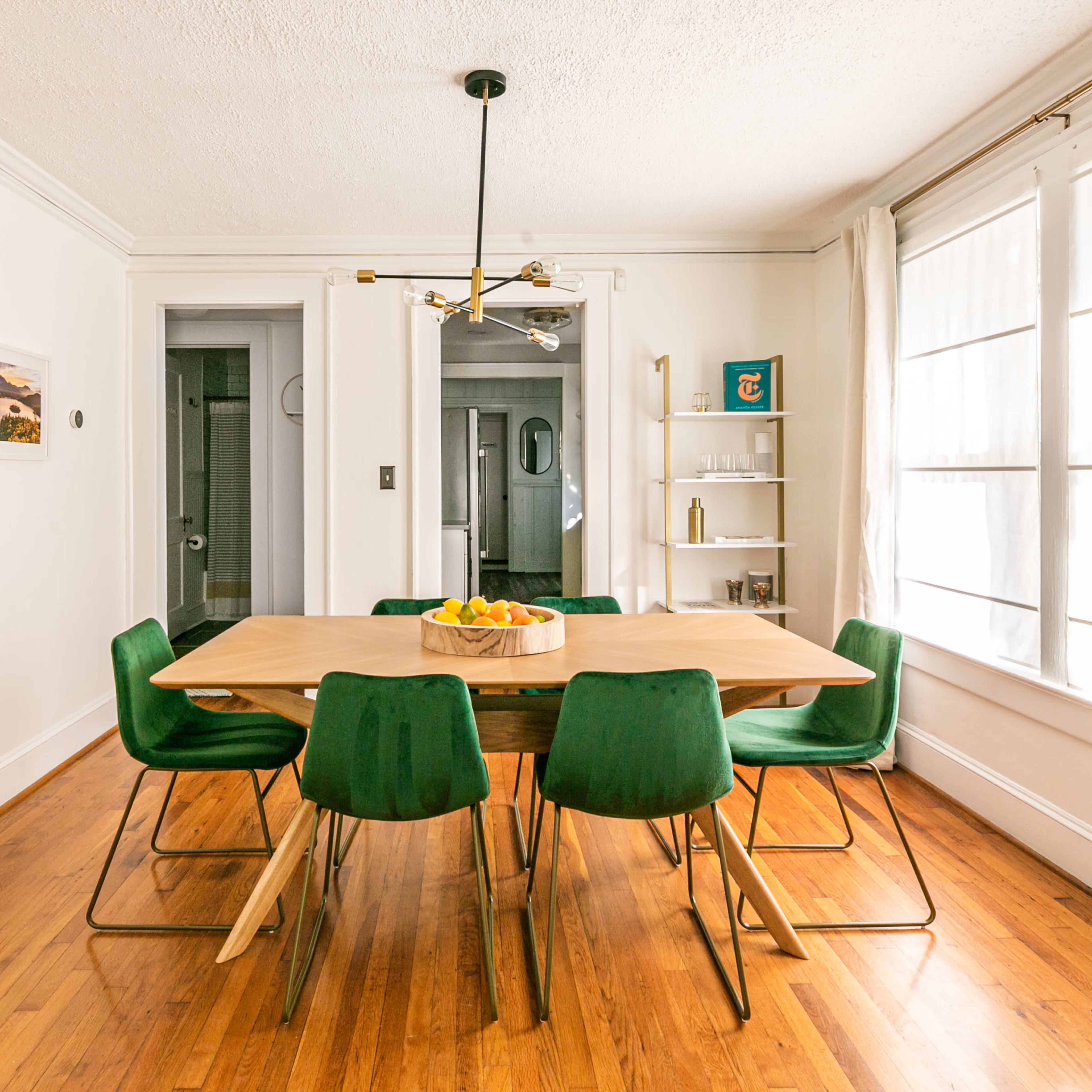 A dining room features a large wooden table surrounded by green chairs, with framed photographs on the walls and large windows letting in natural light.