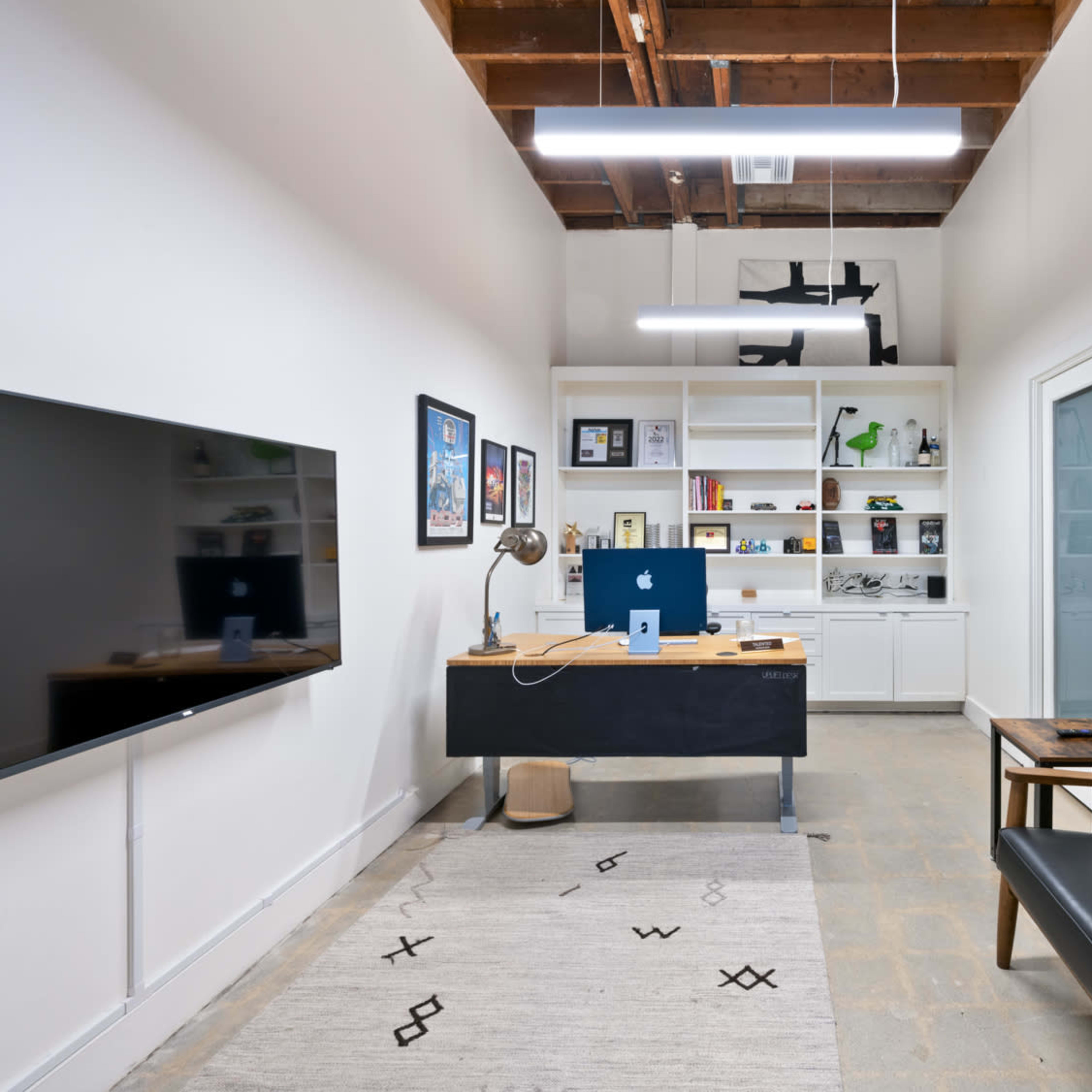A minimalistic office space featuring a wall-mounted TV, a desk with a computer, bookshelves, and a black sofa against the wall.