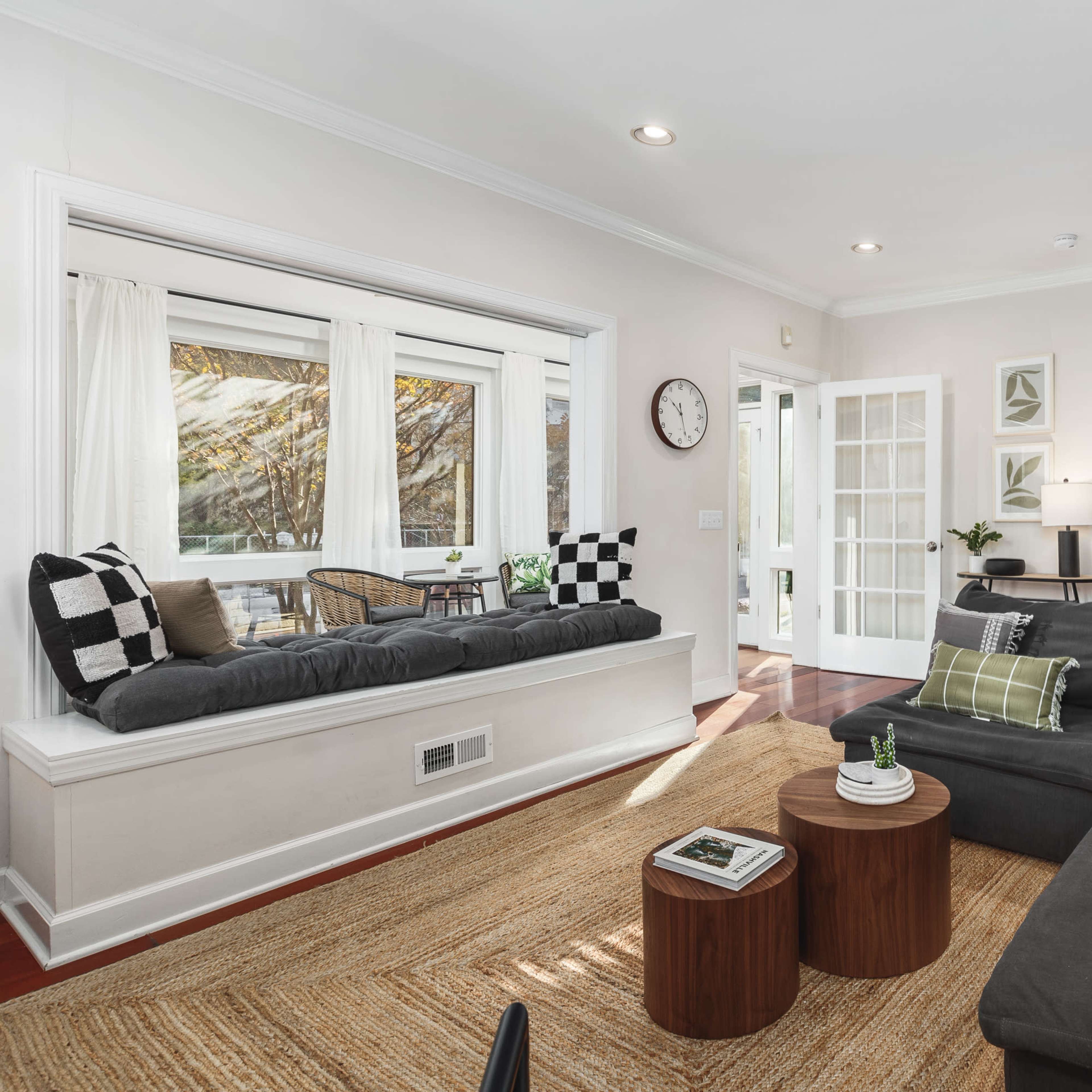 A cozy living room features a sectional sofa, a bay window with seating, two wooden side tables, and a mix of decorative pillows.
