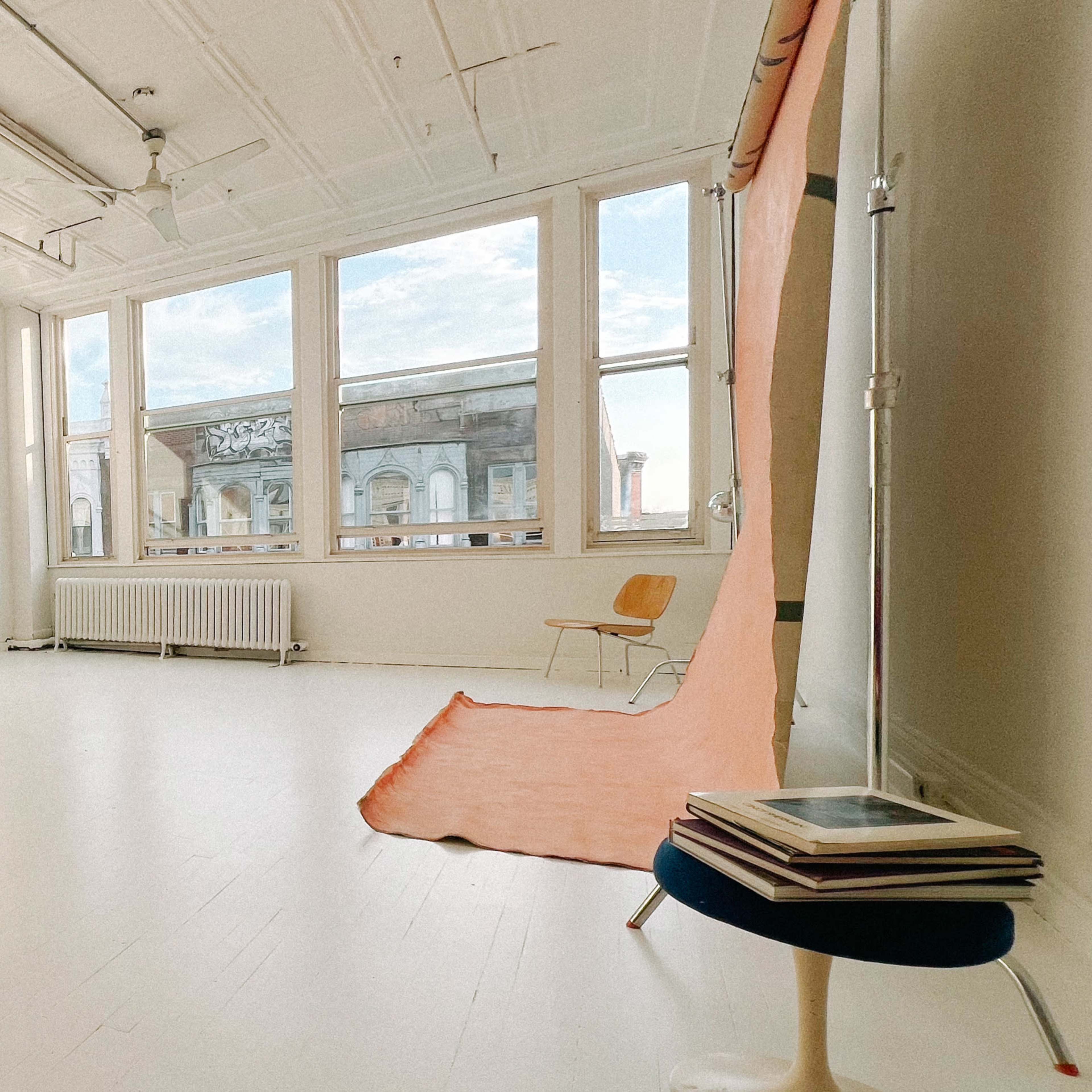 A spacious, well-lit room featuring large windows, a mirror, a chair, and a rolled-out orange backdrop on the floor.