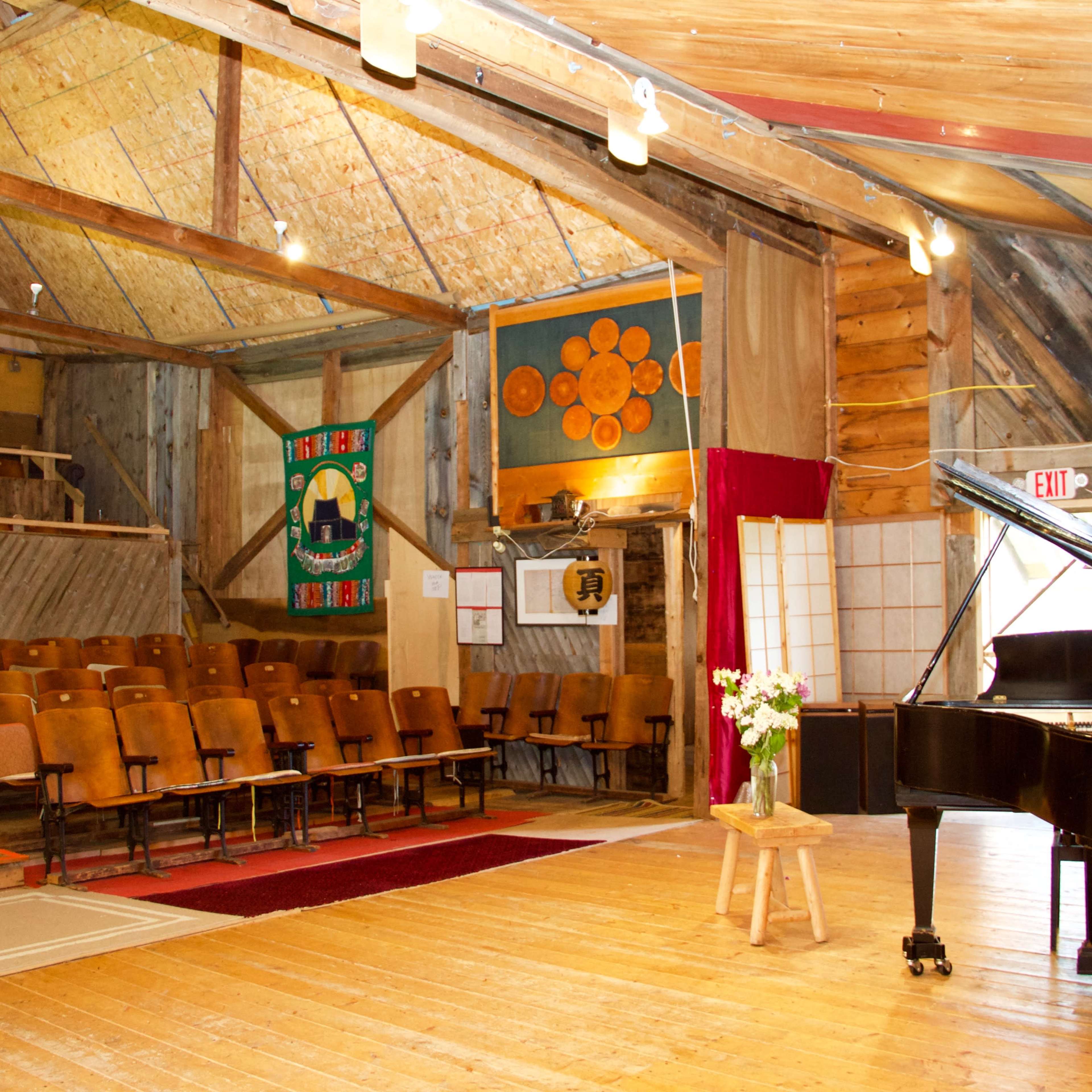 The interior of a rustic hall features a grand piano, wooden floors, rows of orange padded chairs, and colorful banners on the walls.