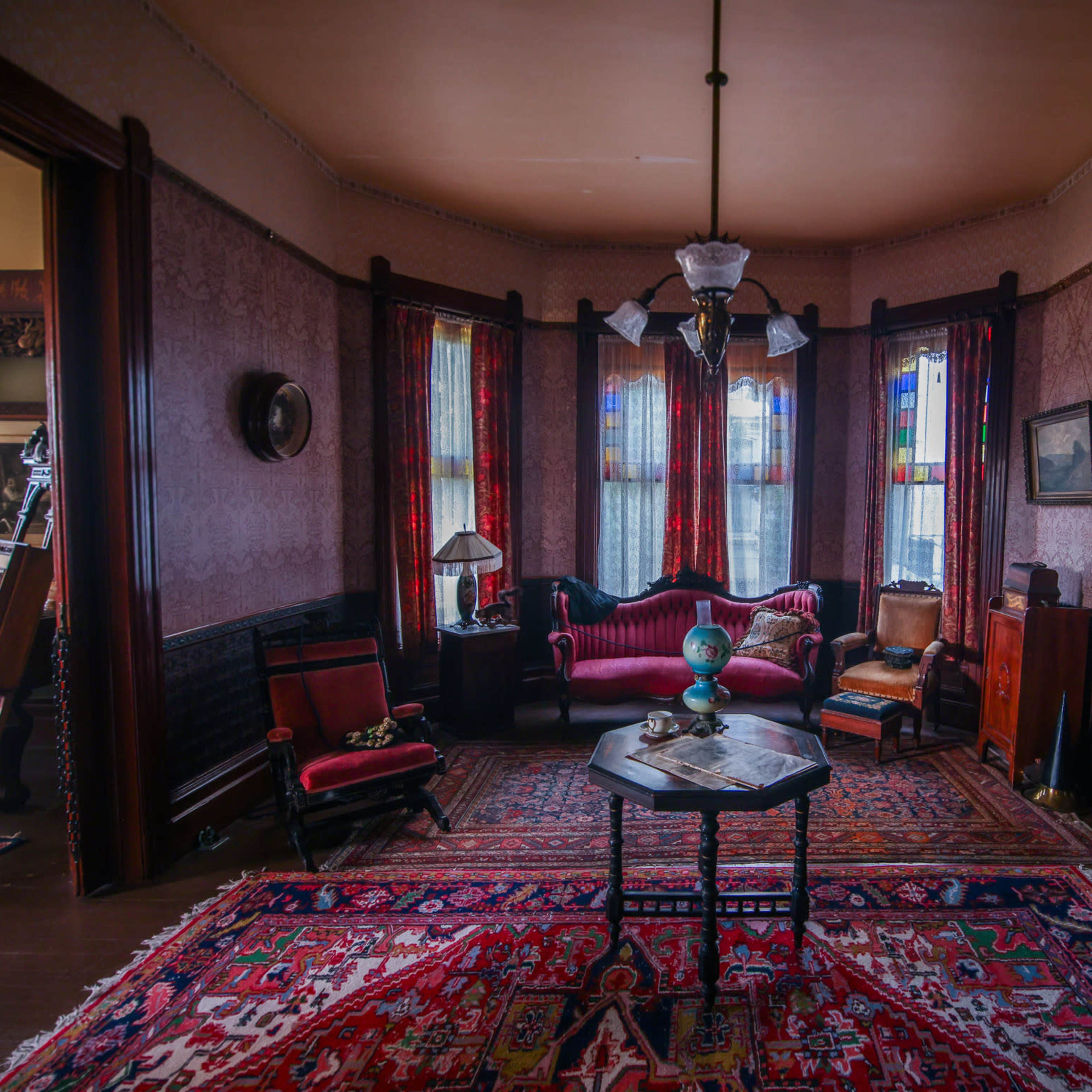 A vintage parlor room with a red patterned rug, a chandelier, stained glass windows, and various pieces of antique furniture.