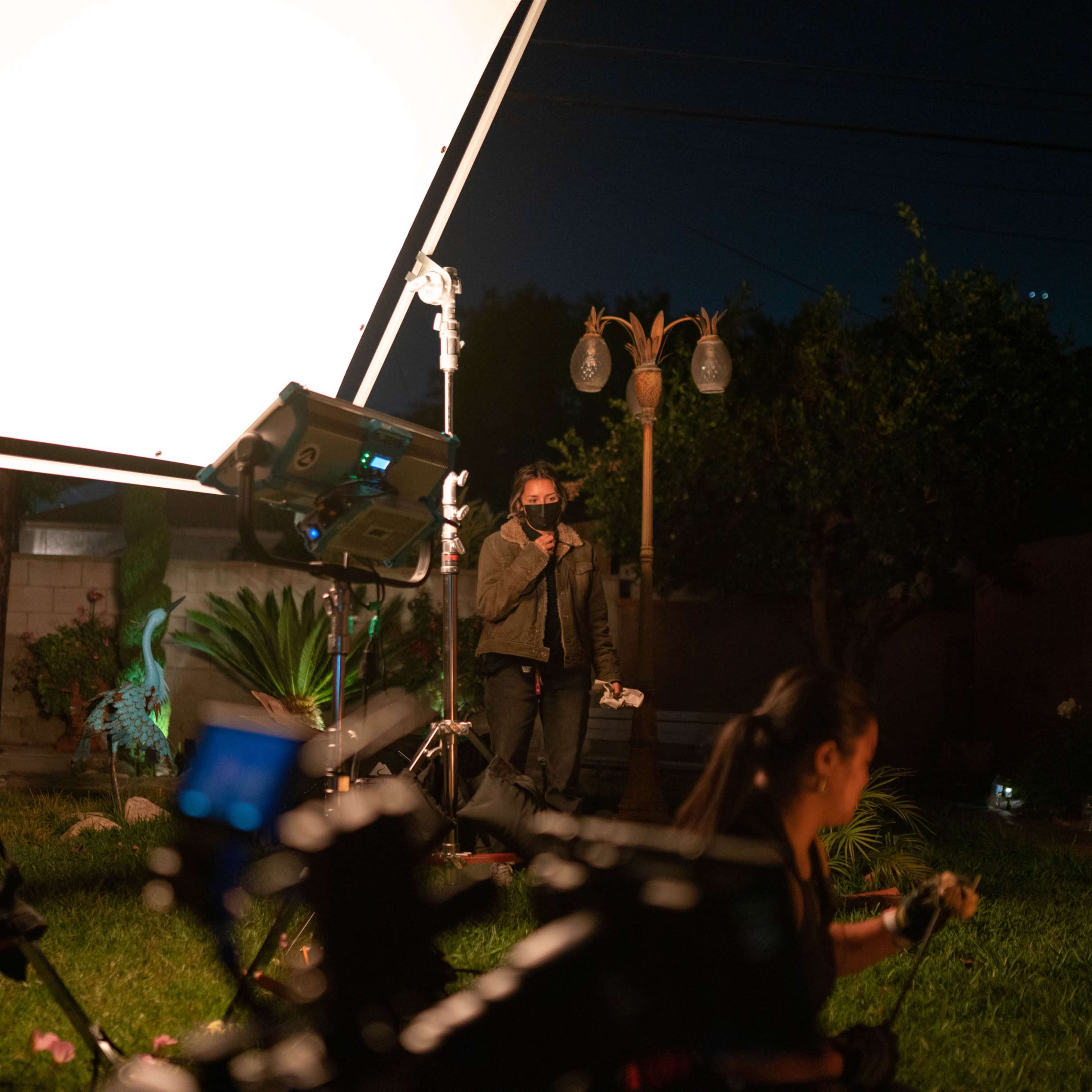 A person wearing a mask stands in a well-lit backyard while film equipment is set up nearby.