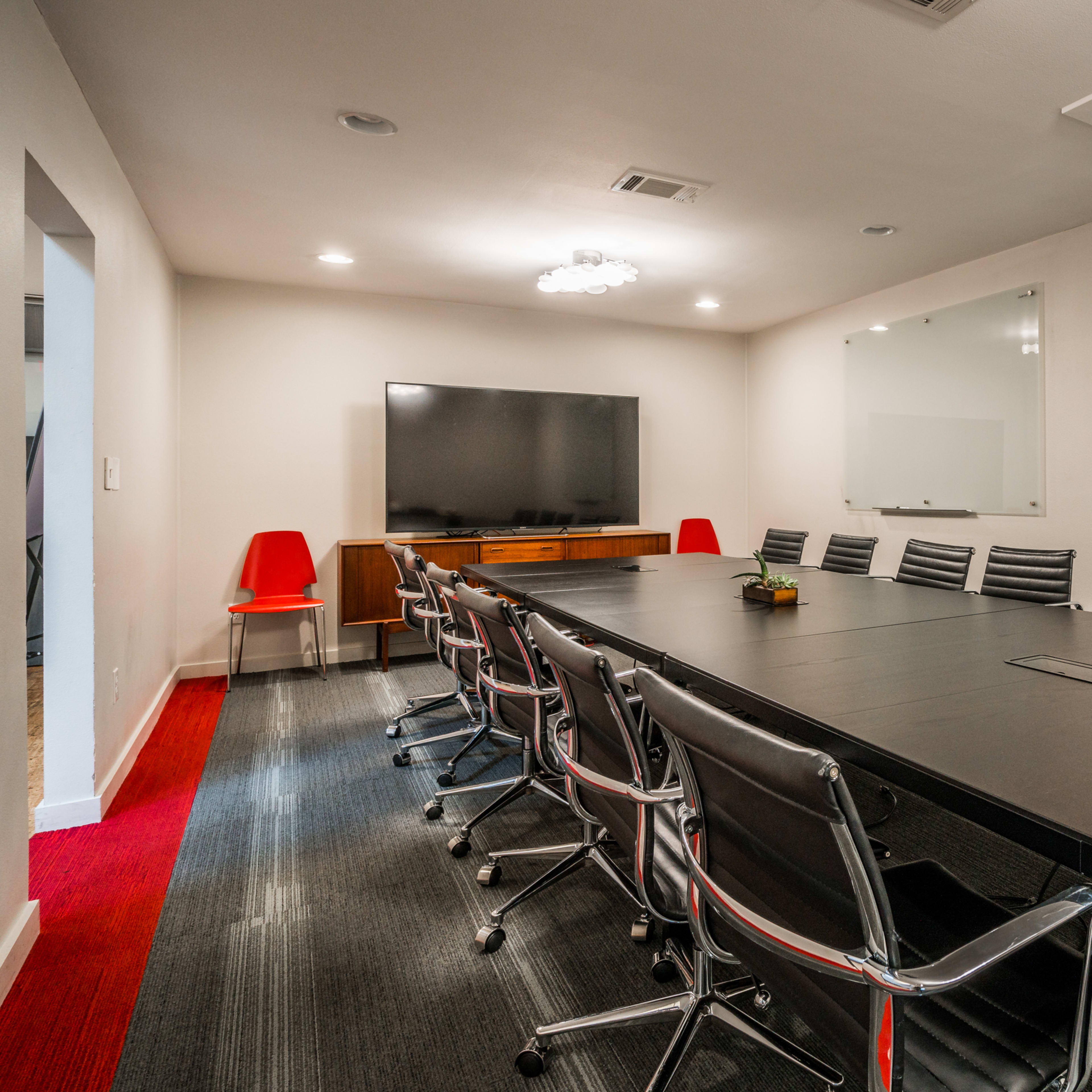 A modern conference room with a long black table, black rolling chairs, a large television screen on one wall, and whiteboard panels on the opposite wall.