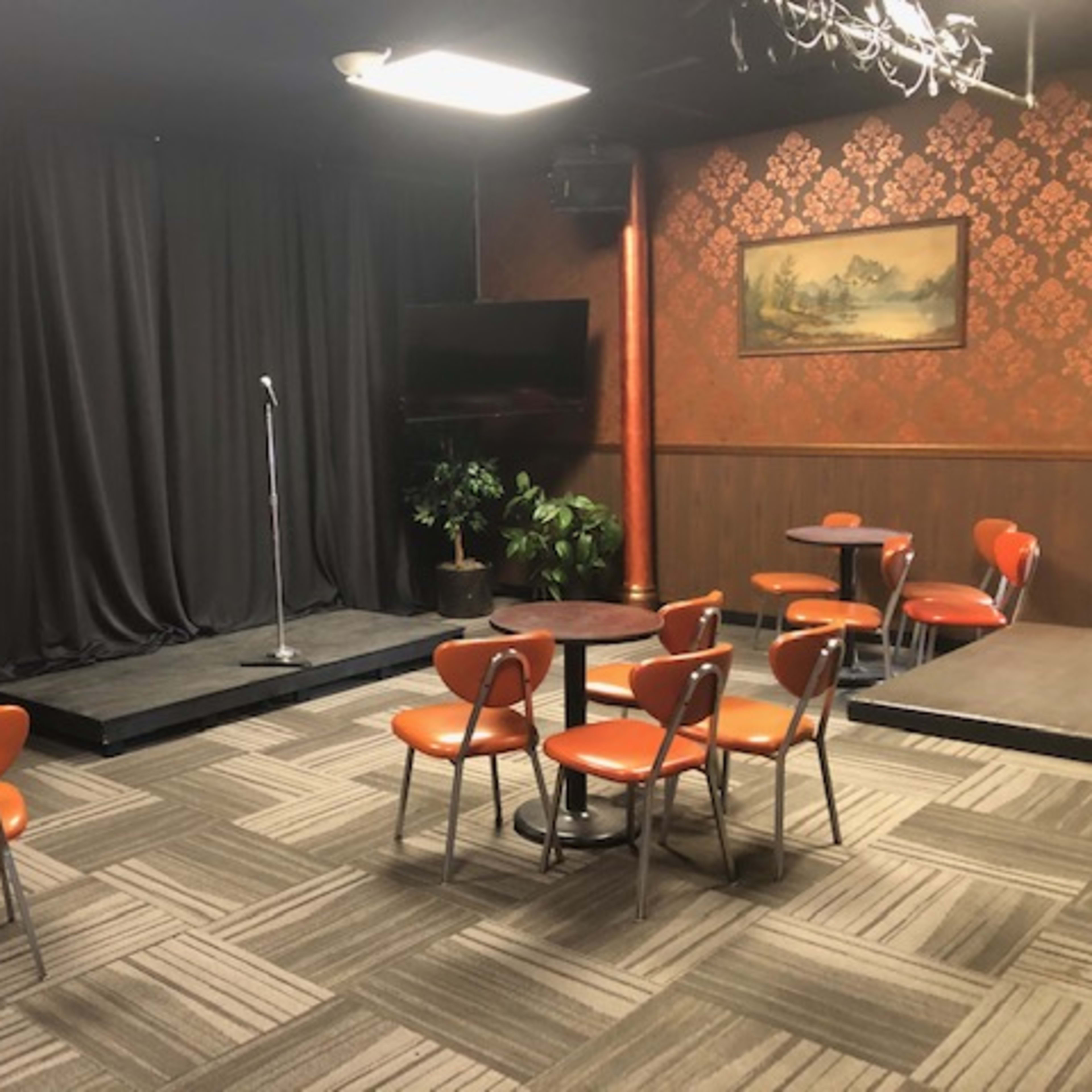 A small performance stage with a microphone, surrounded by several orange chairs and small tables in a dimly lit room with a decorative backdrop.