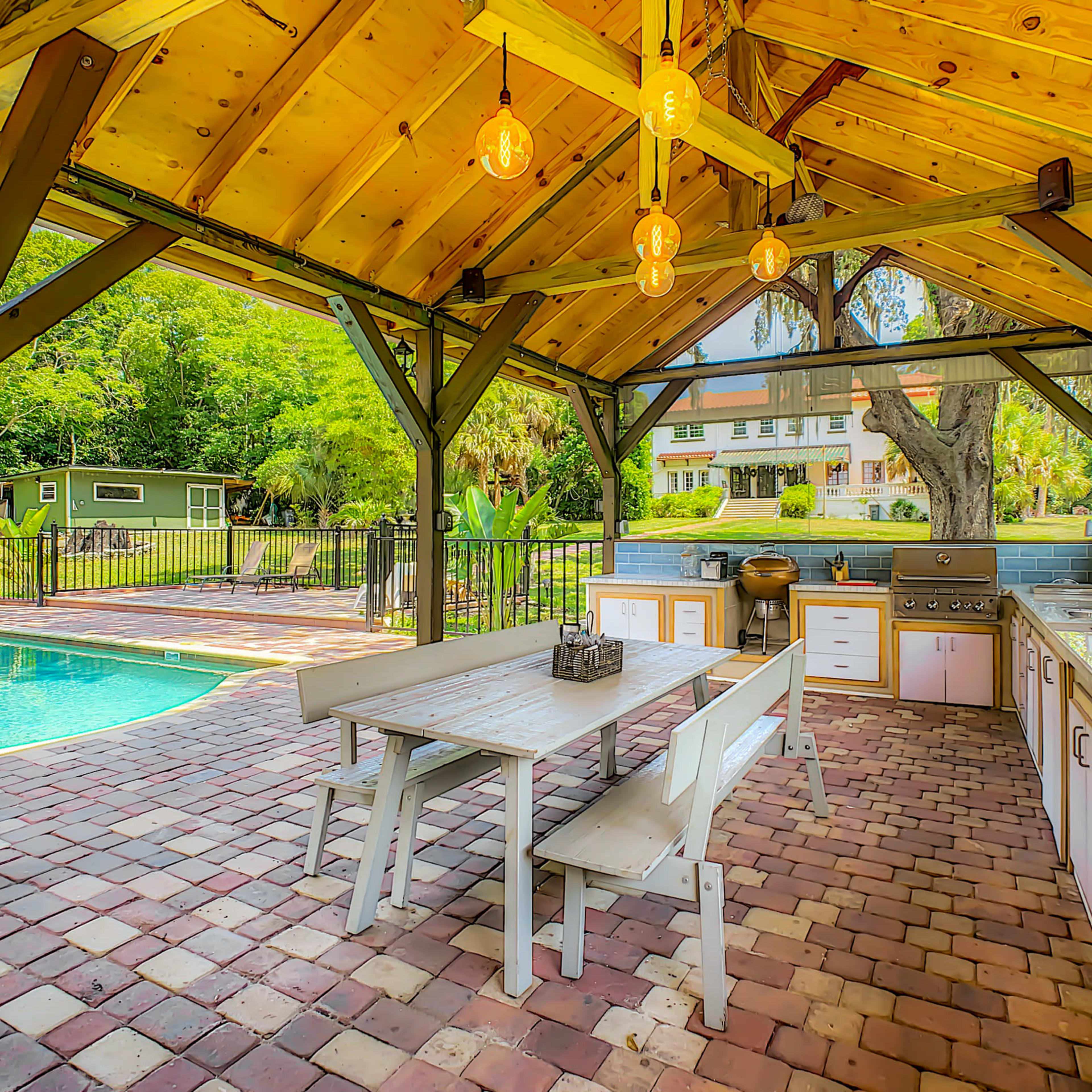 An outdoor kitchen area with a wooden pavilion, a large dining table, and a swimming pool visible in the background.