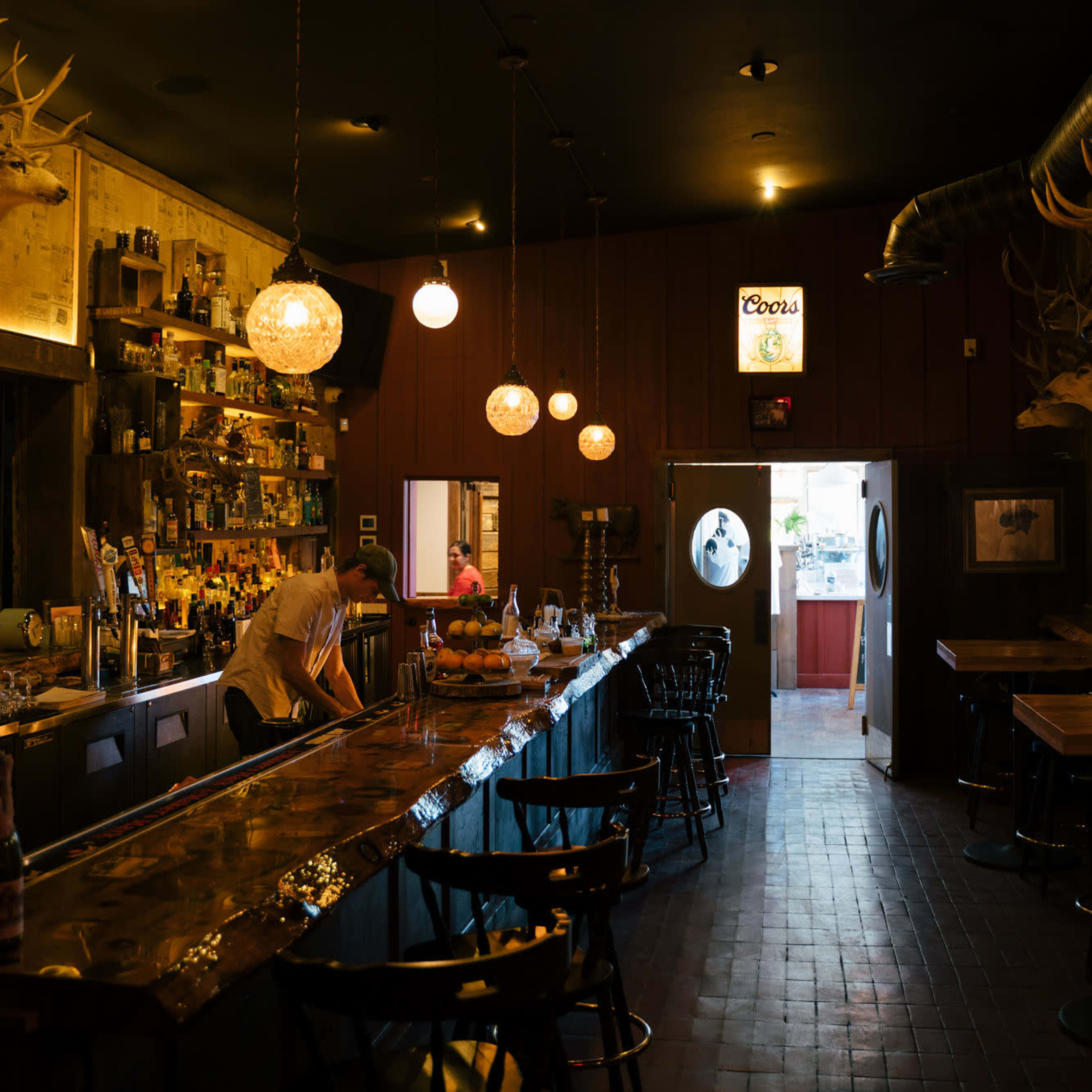 A dimly lit bar features a long counter with several stools, shelves lined with bottles, and round pendant lights hanging from the ceiling.