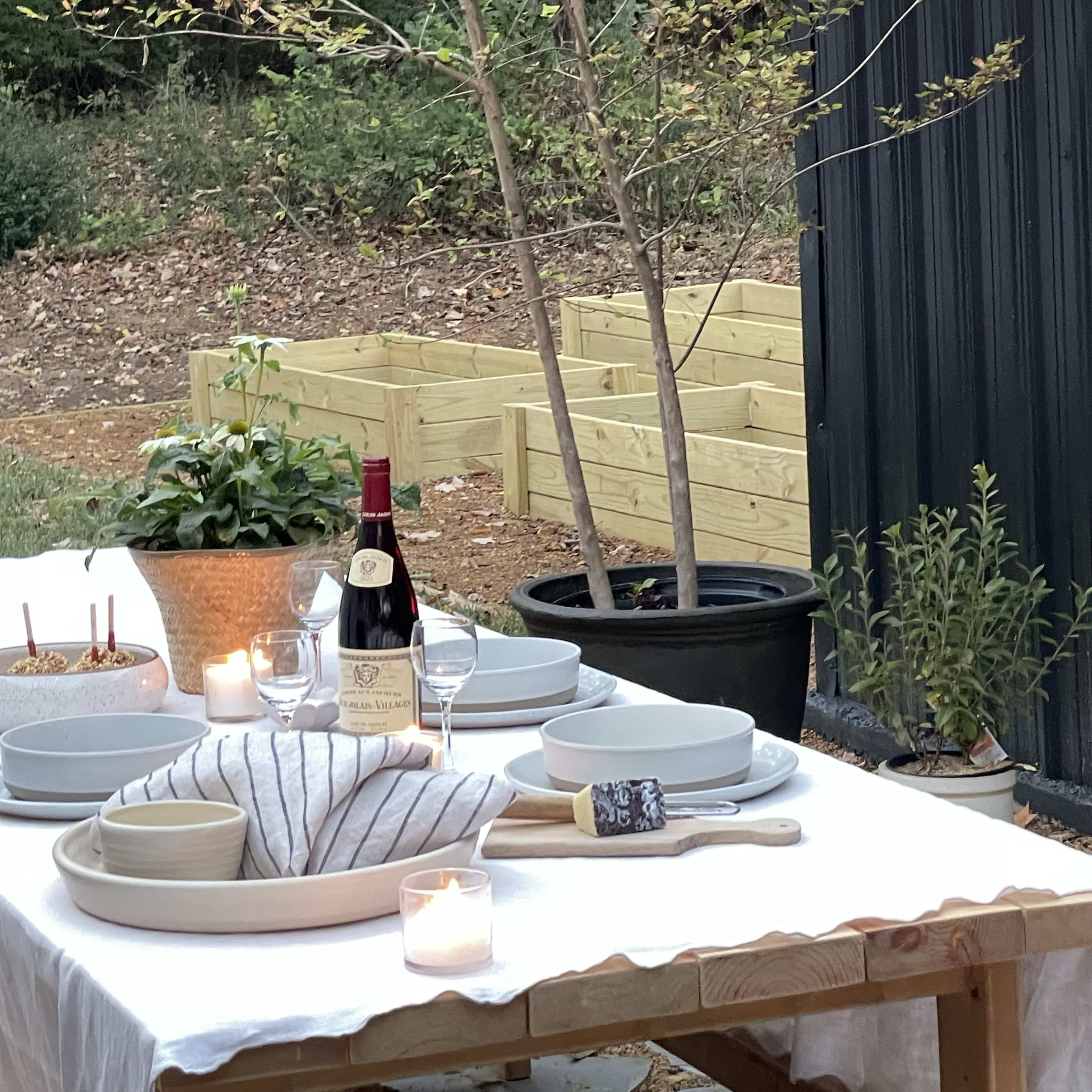 An outdoor dining table is set with plates, glasses, and a bottle of wine, beside potted plants and a garden bed.