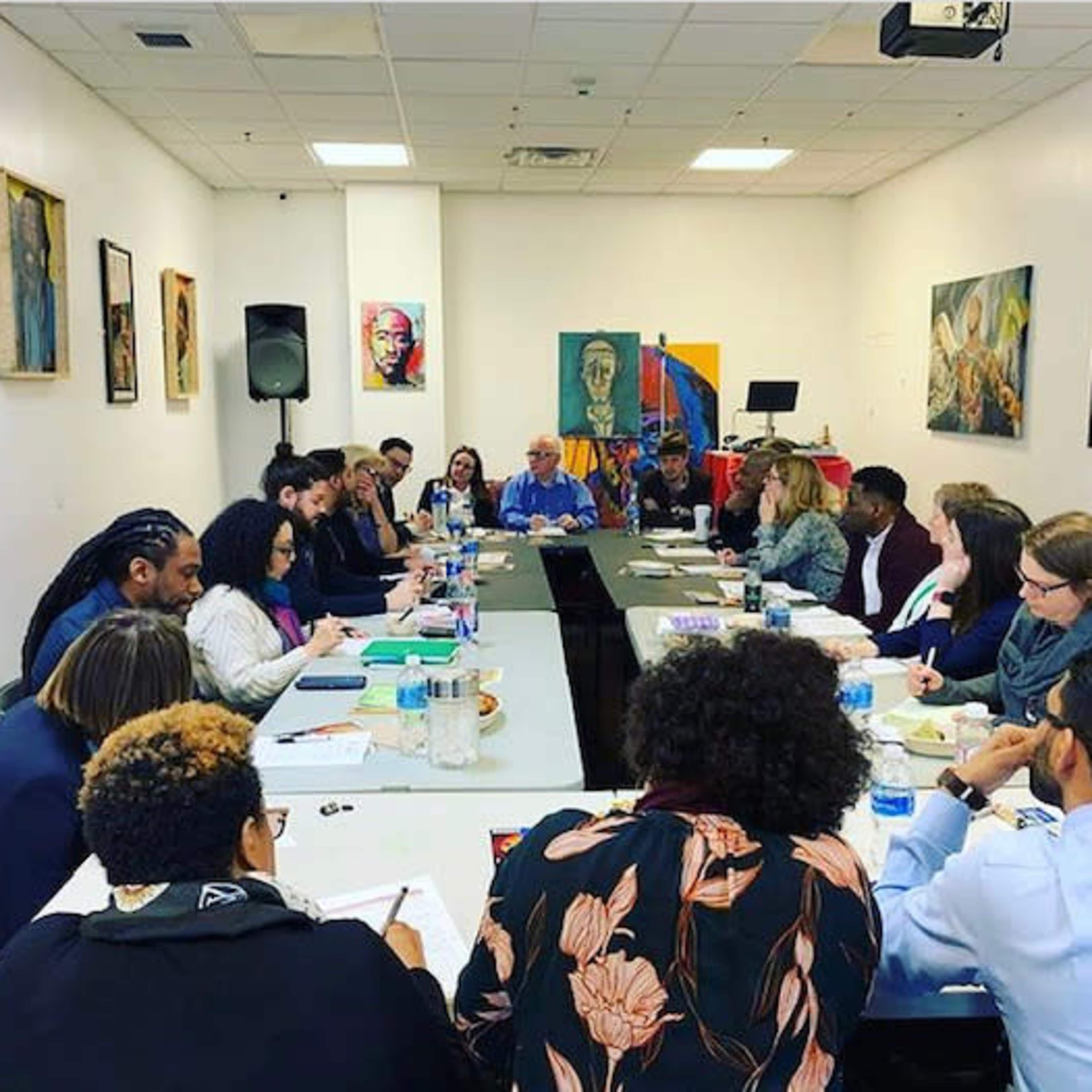 A diverse group of people is seated around a long table in a well-lit conference room, engaged in discussion while various artworks adorn the walls.