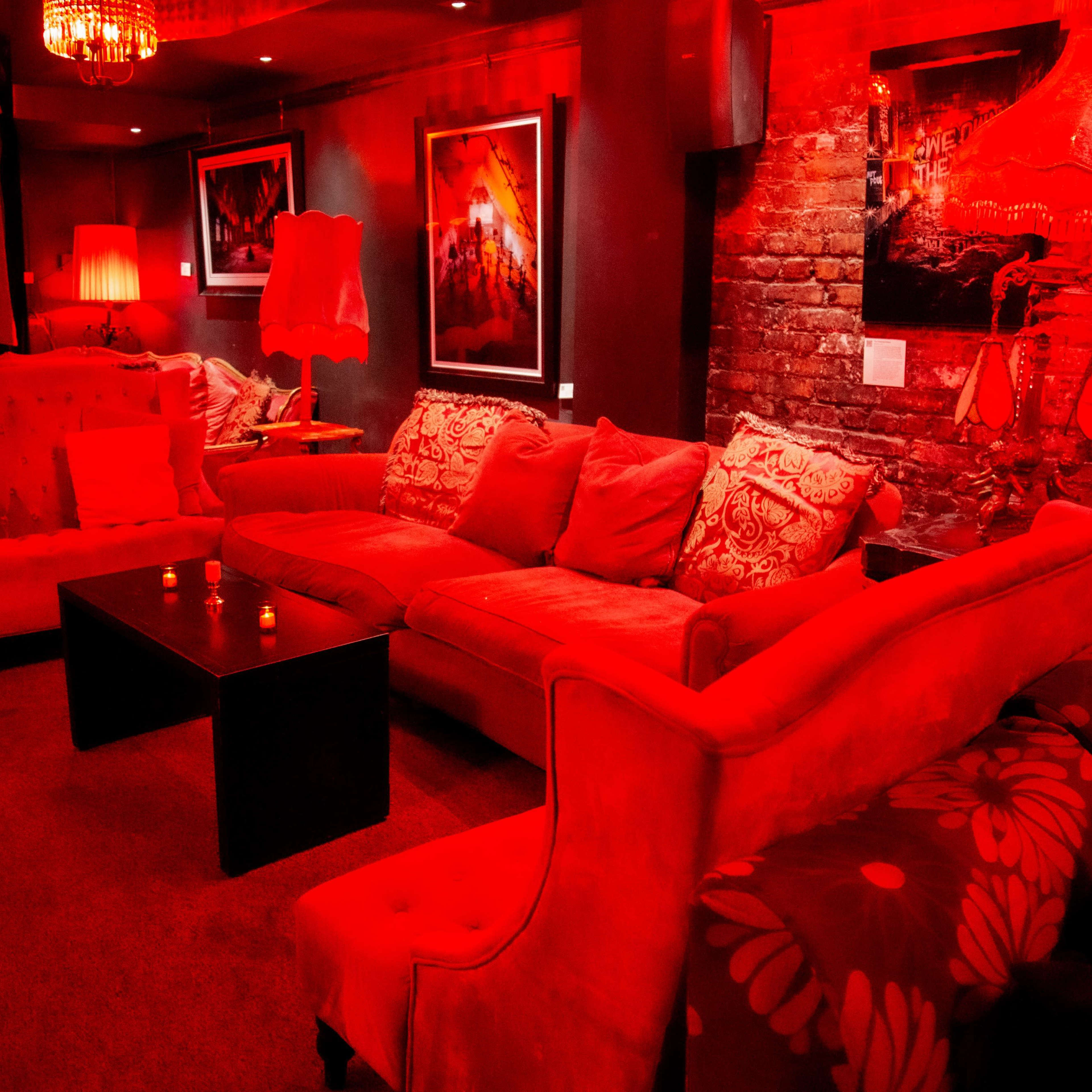 A dimly lit lounge features red sofas, a coffee table, and artwork on the walls against a brick backdrop.