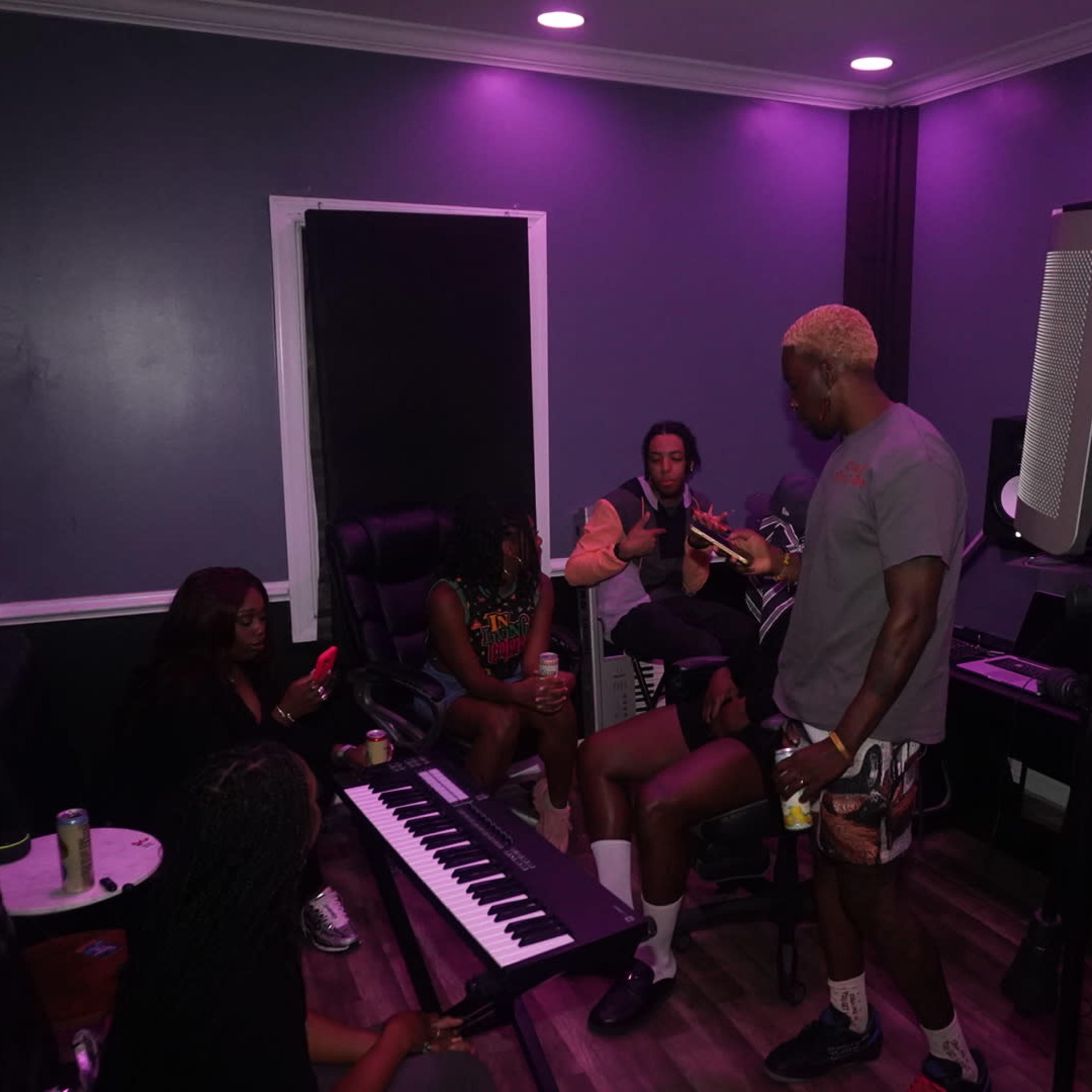A group of five people is gathered in a music studio with purple lighting, engaged in conversation while a keyboard and recording equipment are present.