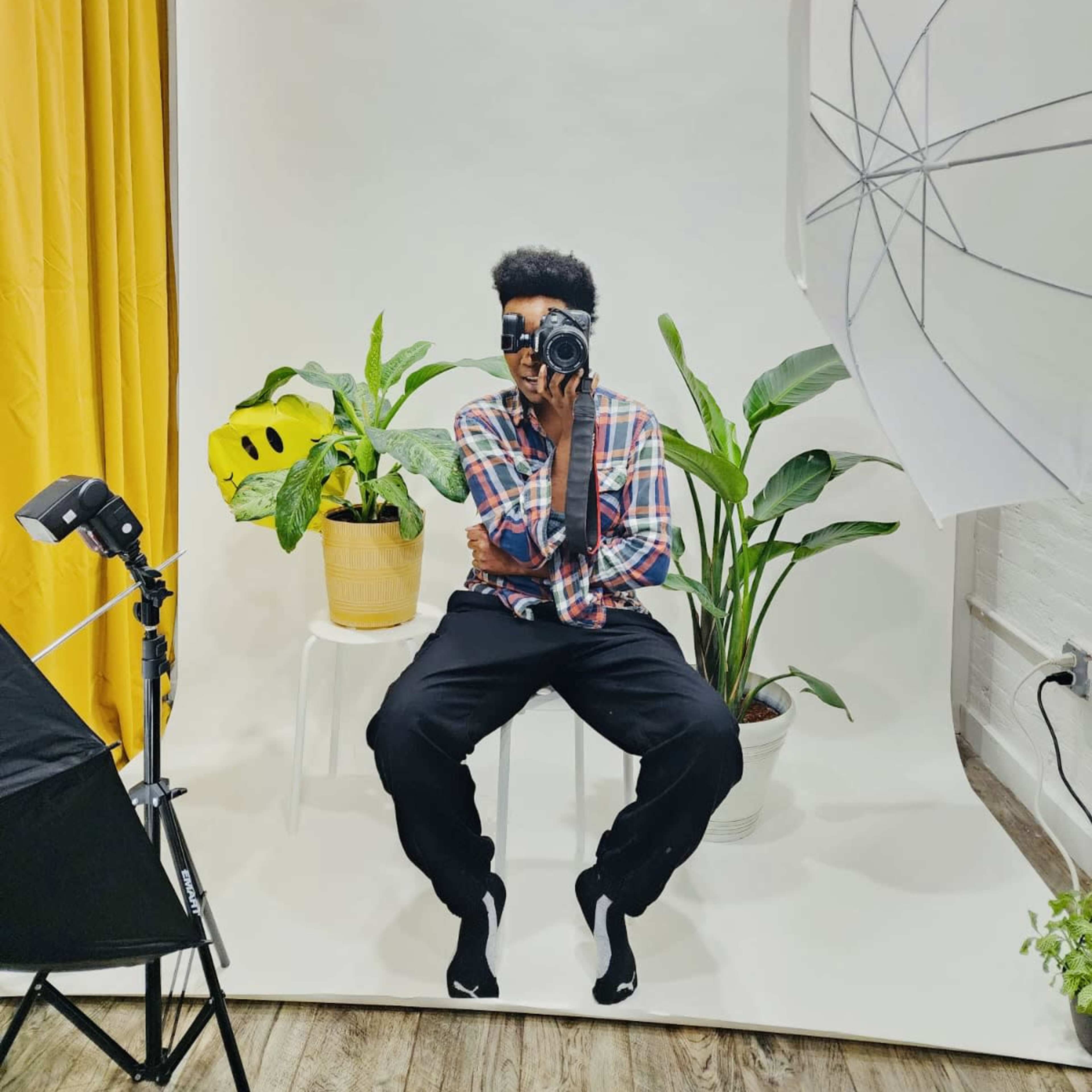 A person is sitting on a white stool in front of a light backdrop, holding a camera, surrounded by potted plants and studio lighting equipment.