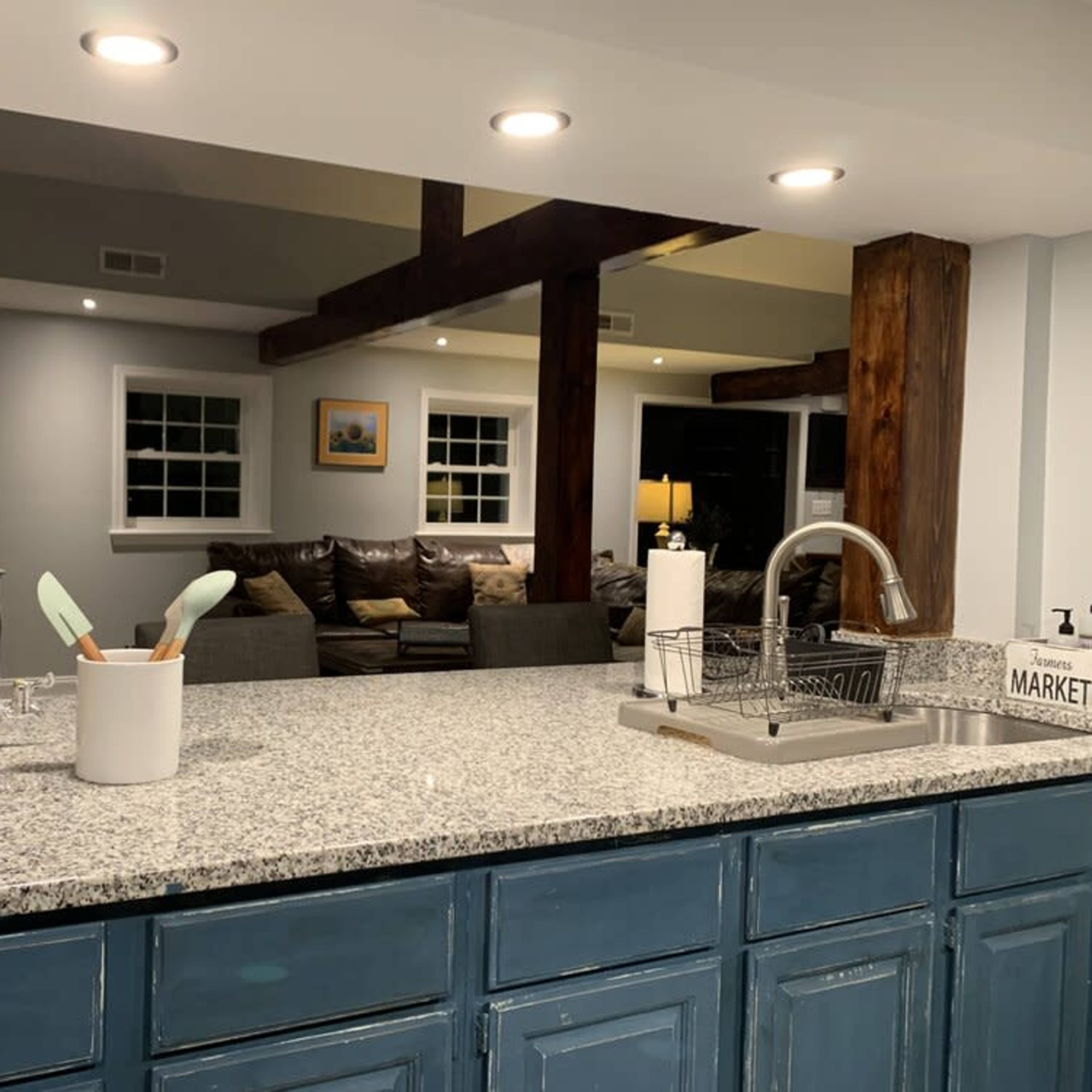 A modern kitchen with a granite countertop, a sink, and a view of a living area with a sofa and windows in the background.