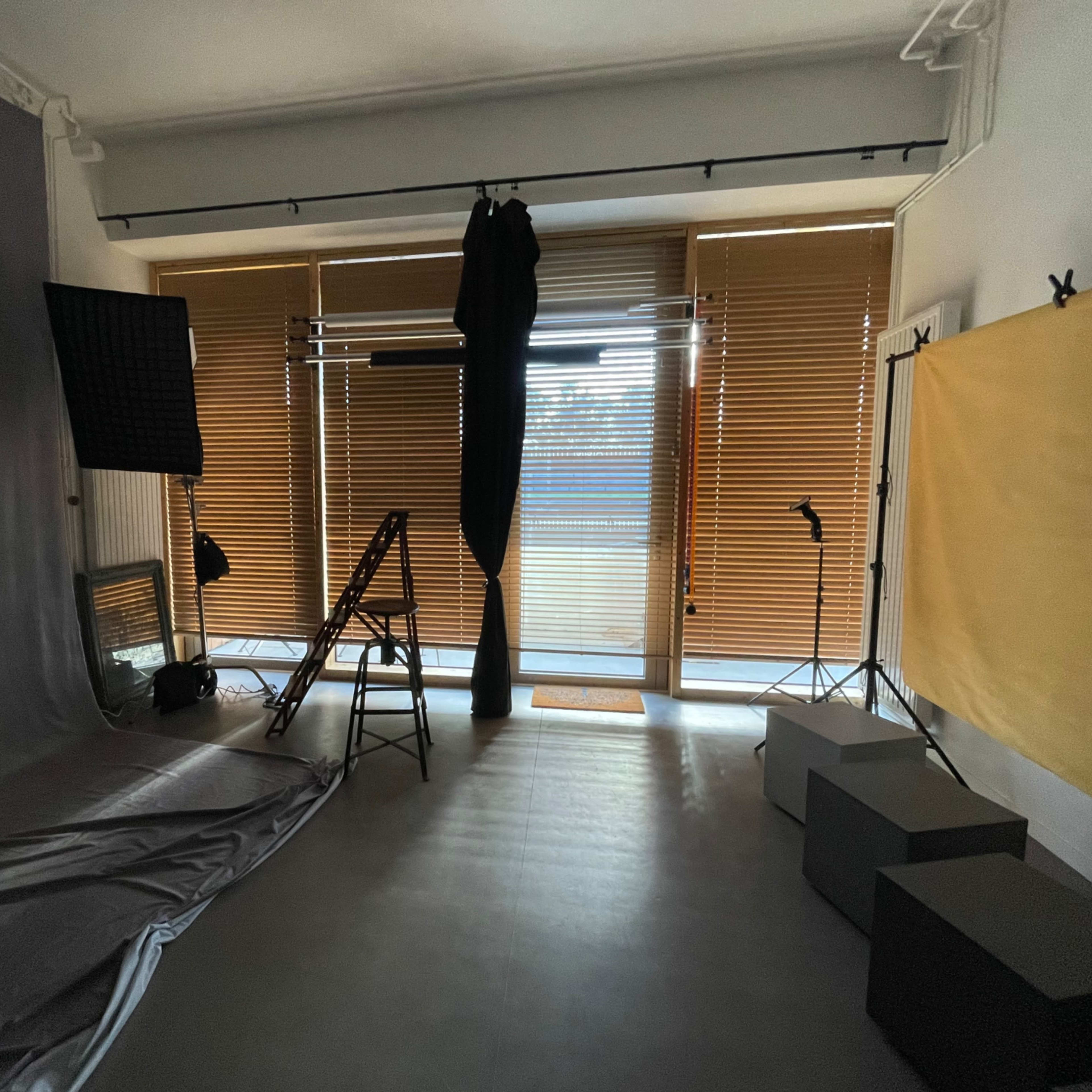 A photography studio with equipment, including softboxes, a backdrop, and a setup for lighting, along with large windows covered by blinds.