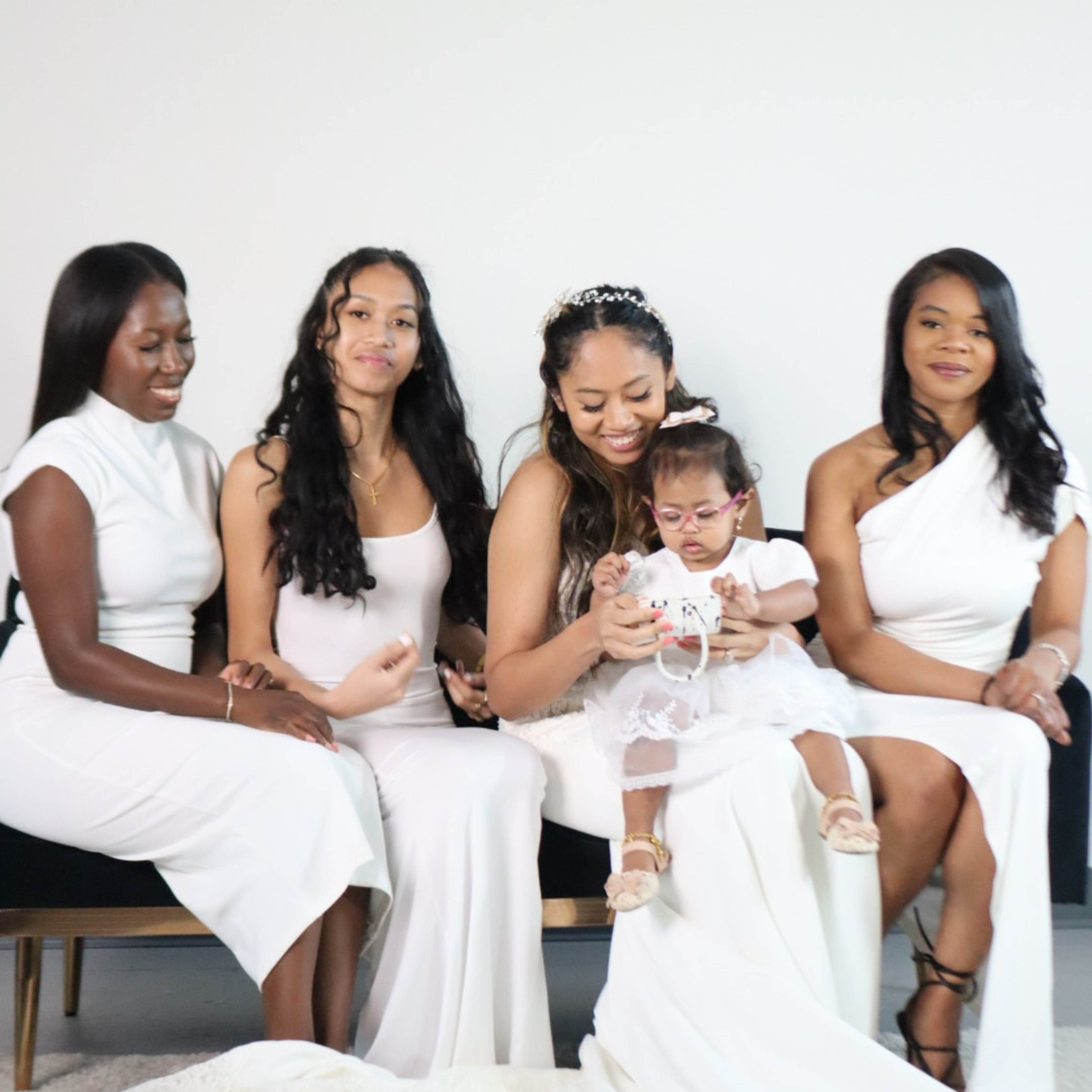 Five women in white dresses sit together, with one woman holding a young child in a white outfit.