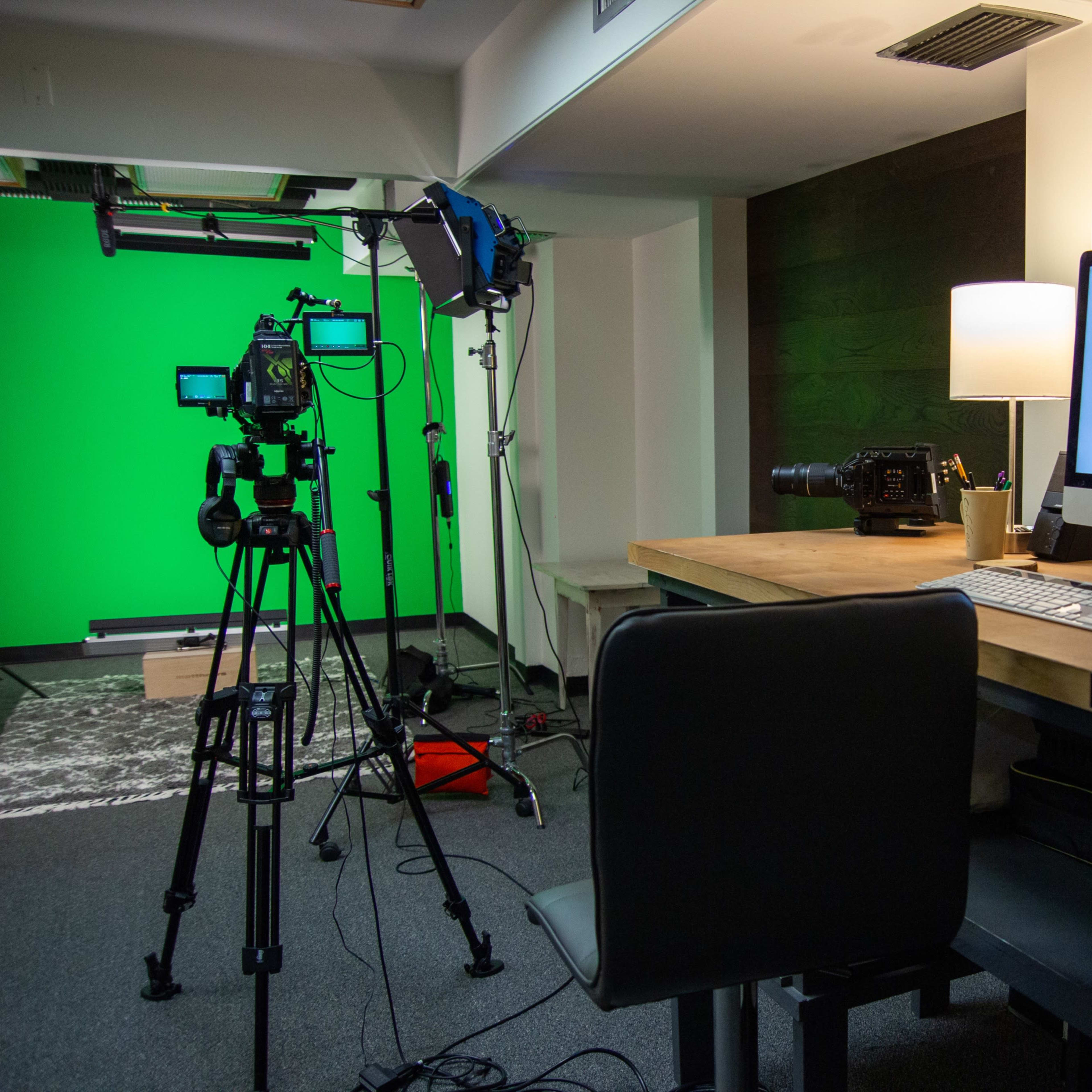 A production studio with a green screen, camera equipment, a computer displaying the logo "DUCK DUCK PRODUCTIONS," and a desk with a chair.