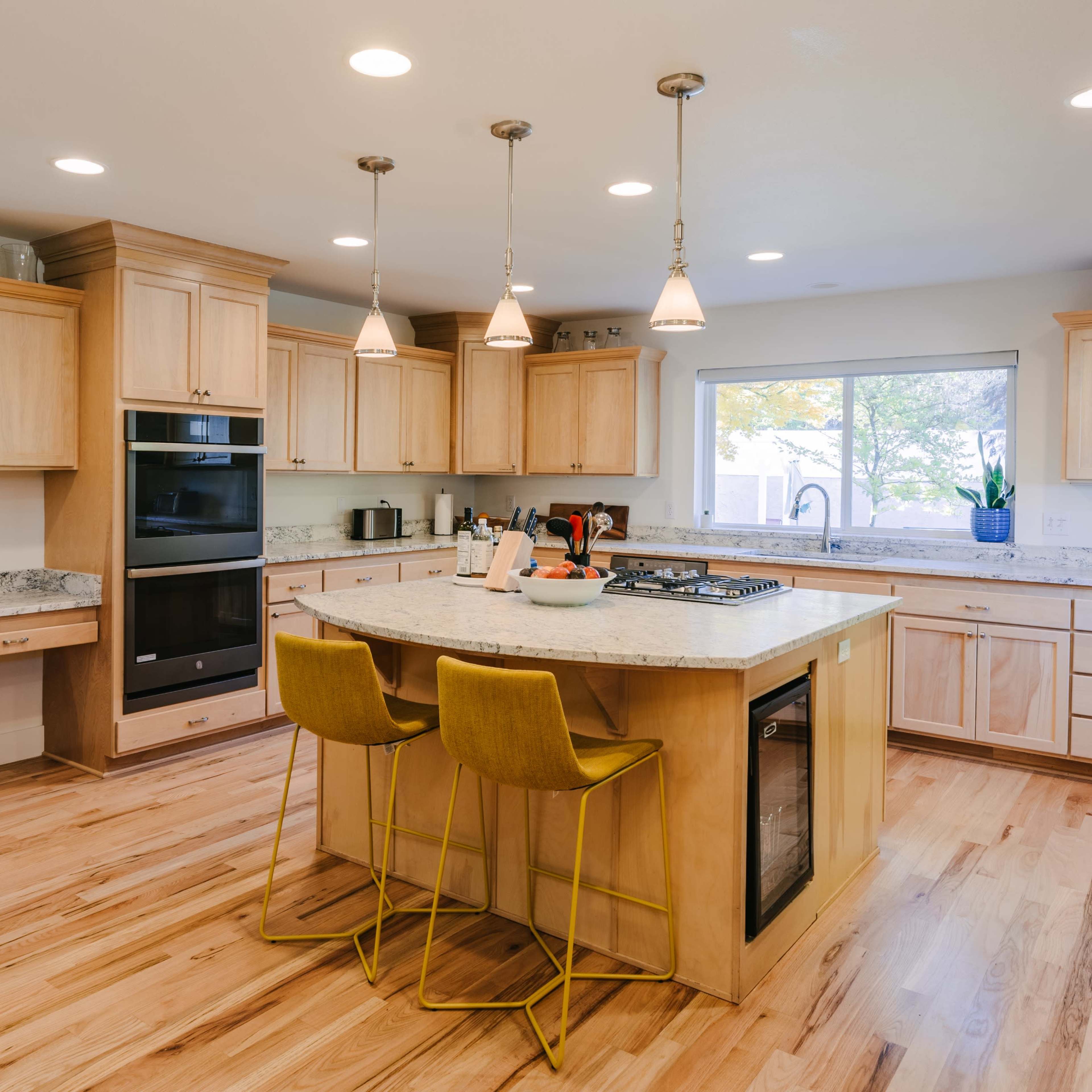 A modern kitchen features light wood cabinets, a central island with three yellow bar stools, and stainless steel appliances.