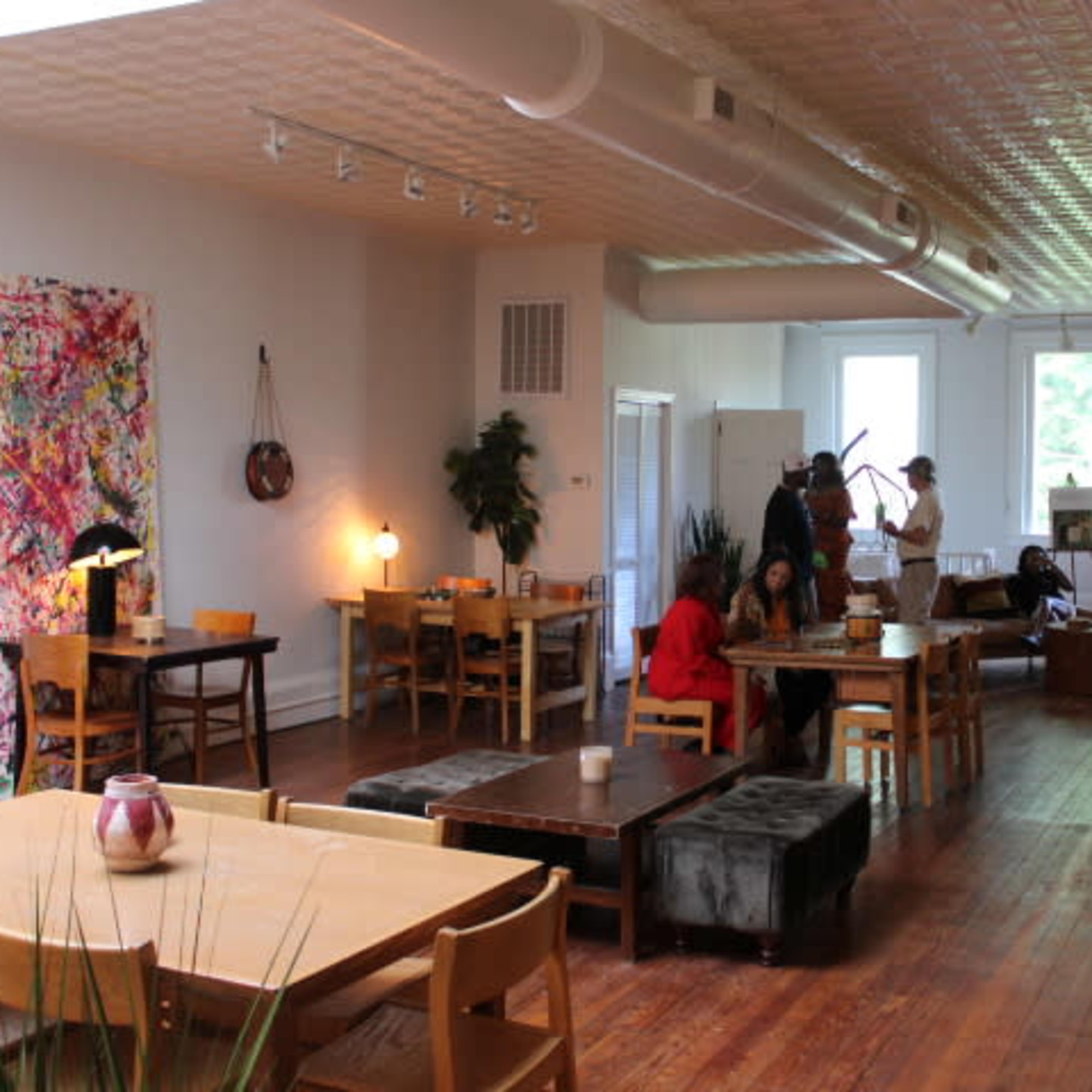 A spacious café with wooden floors and various seating arrangements, including tables and couches, accompanied by art pieces and plants.
