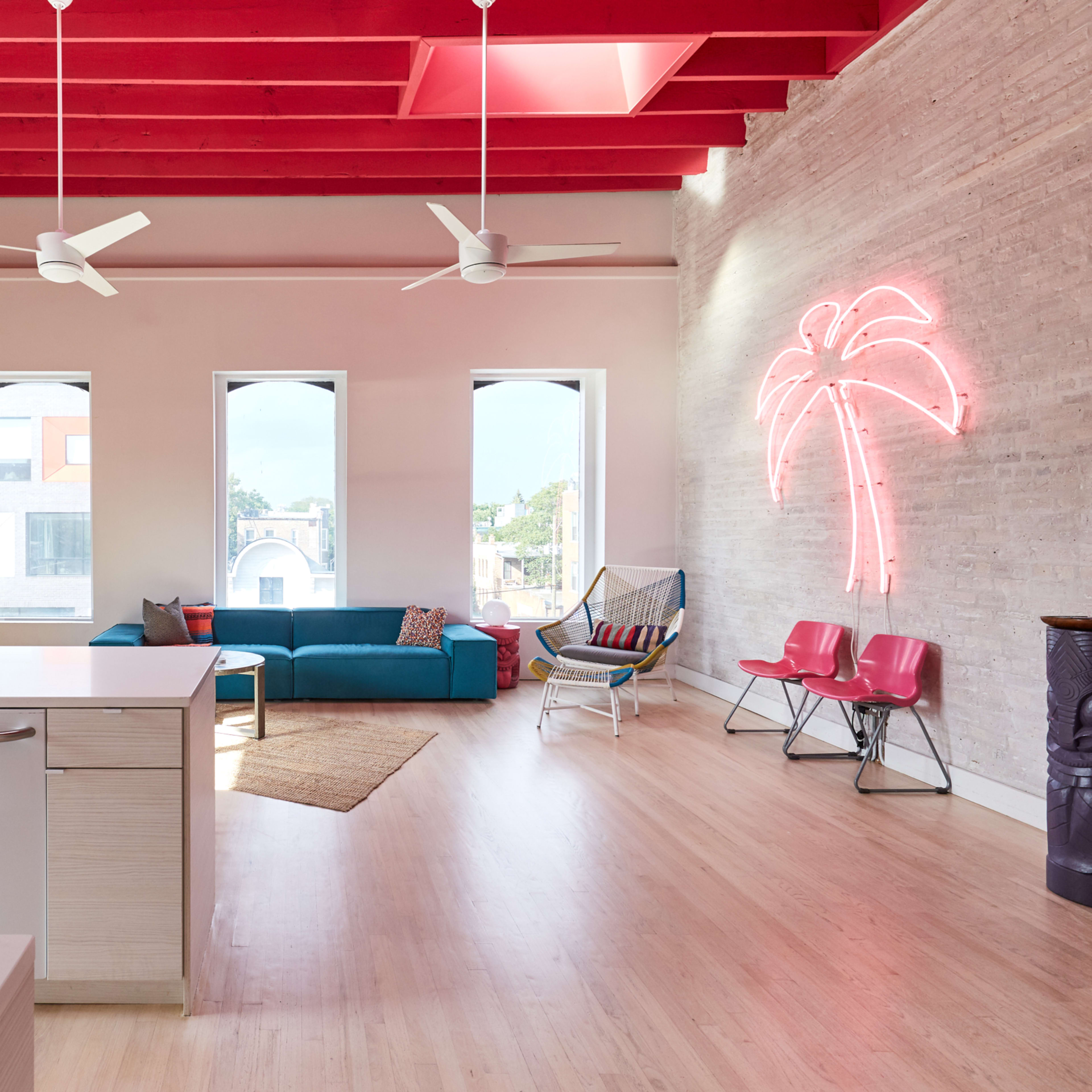 A bright, modern living space with red beams, light wood flooring, and distinct decor elements including a pink shark sculpture and a neon palm tree.
