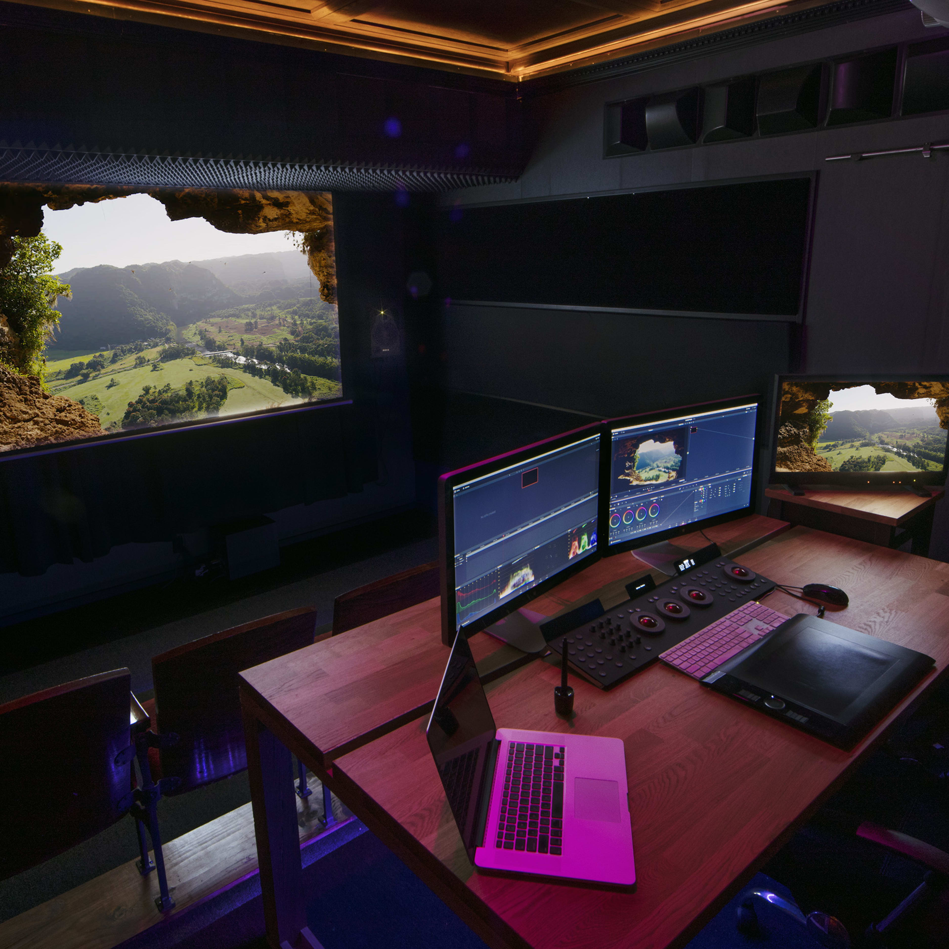 A modern editing studio with two computer monitors and a laptop, featuring a view of a scenic landscape through a window-like opening in a stone wall.