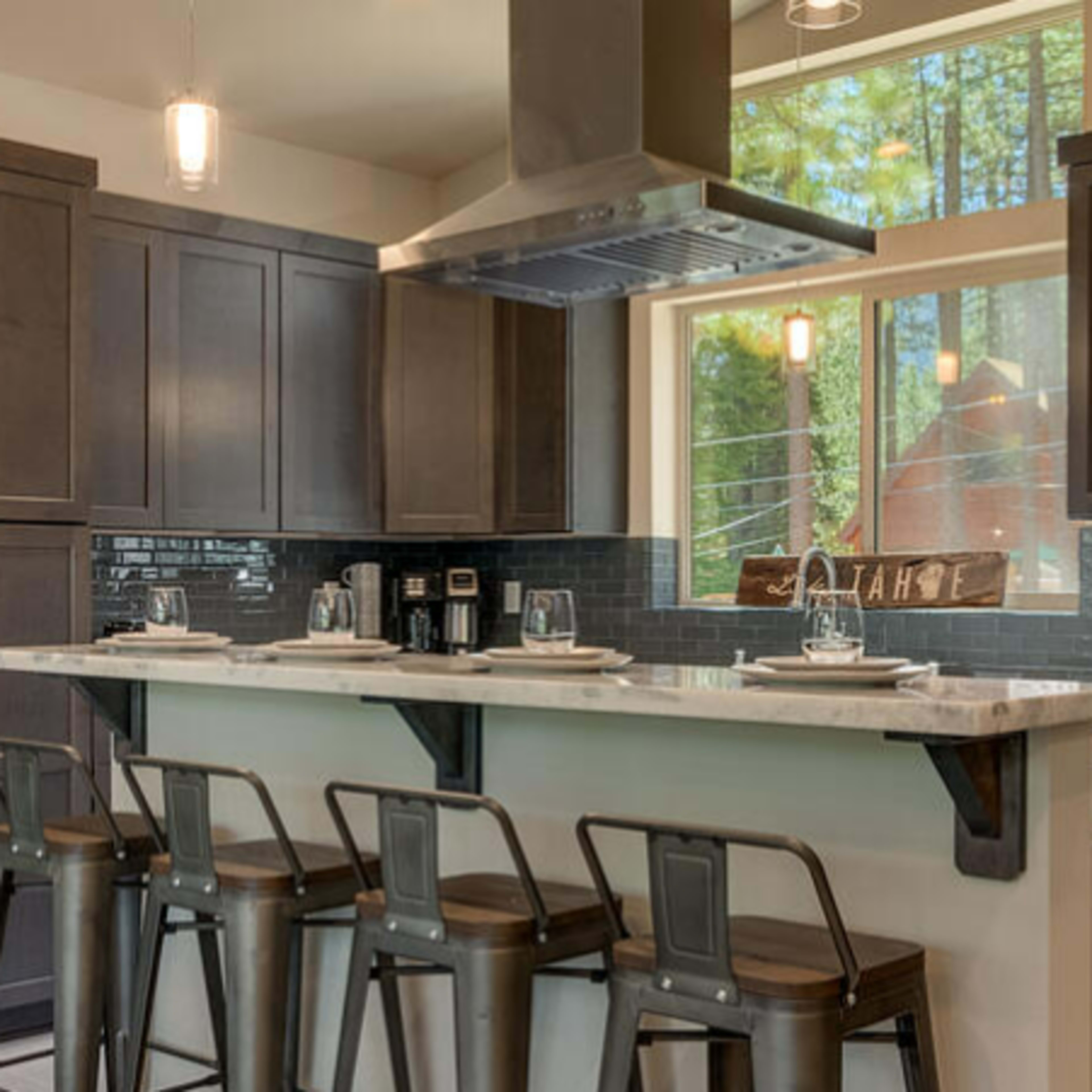 A modern kitchen with dark cabinetry, a central island with bar stools, and a large window overlooking greenery.