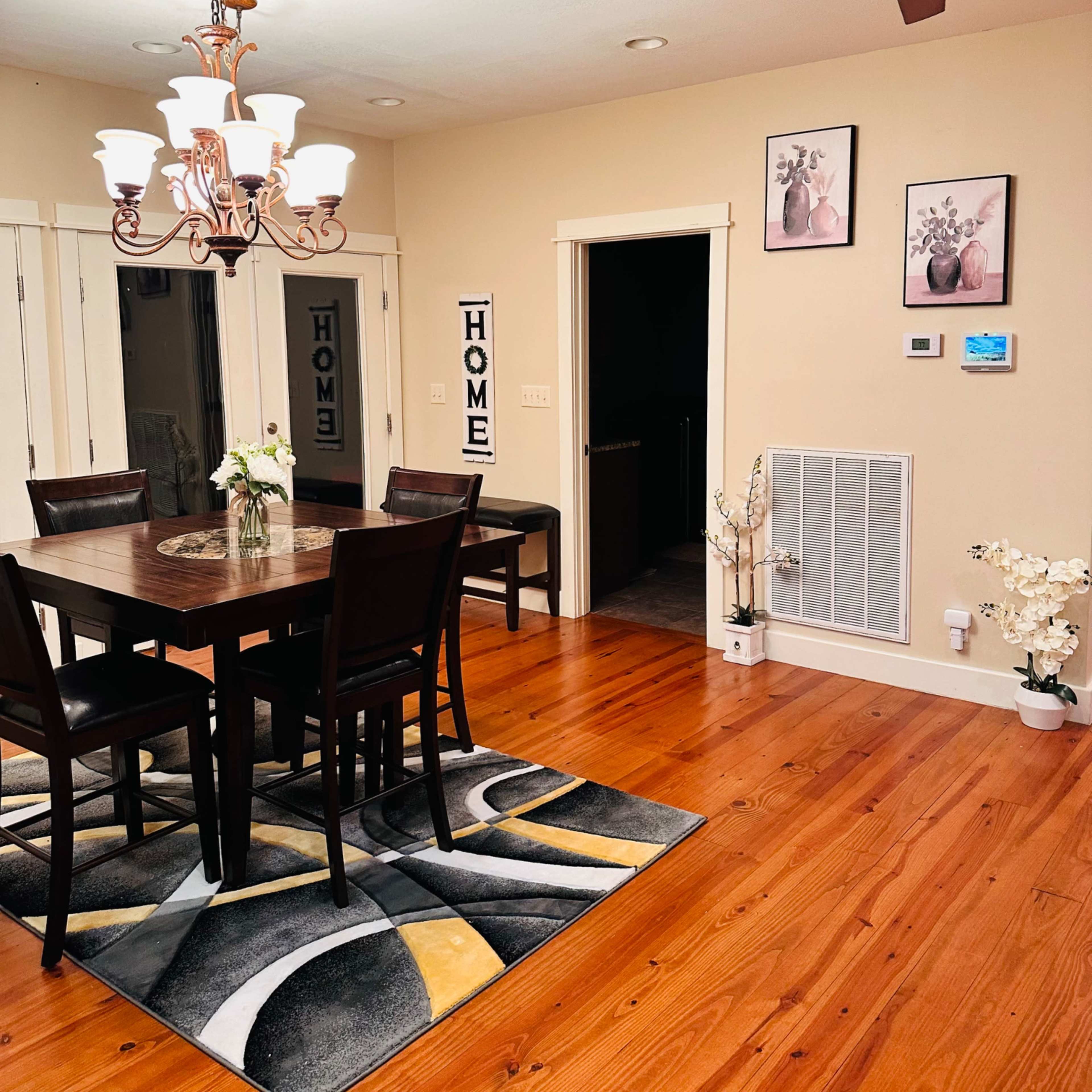 A dining room features a wooden table surrounded by chairs, a chandelier overhead, and decorative elements like wall art and a patterned rug.