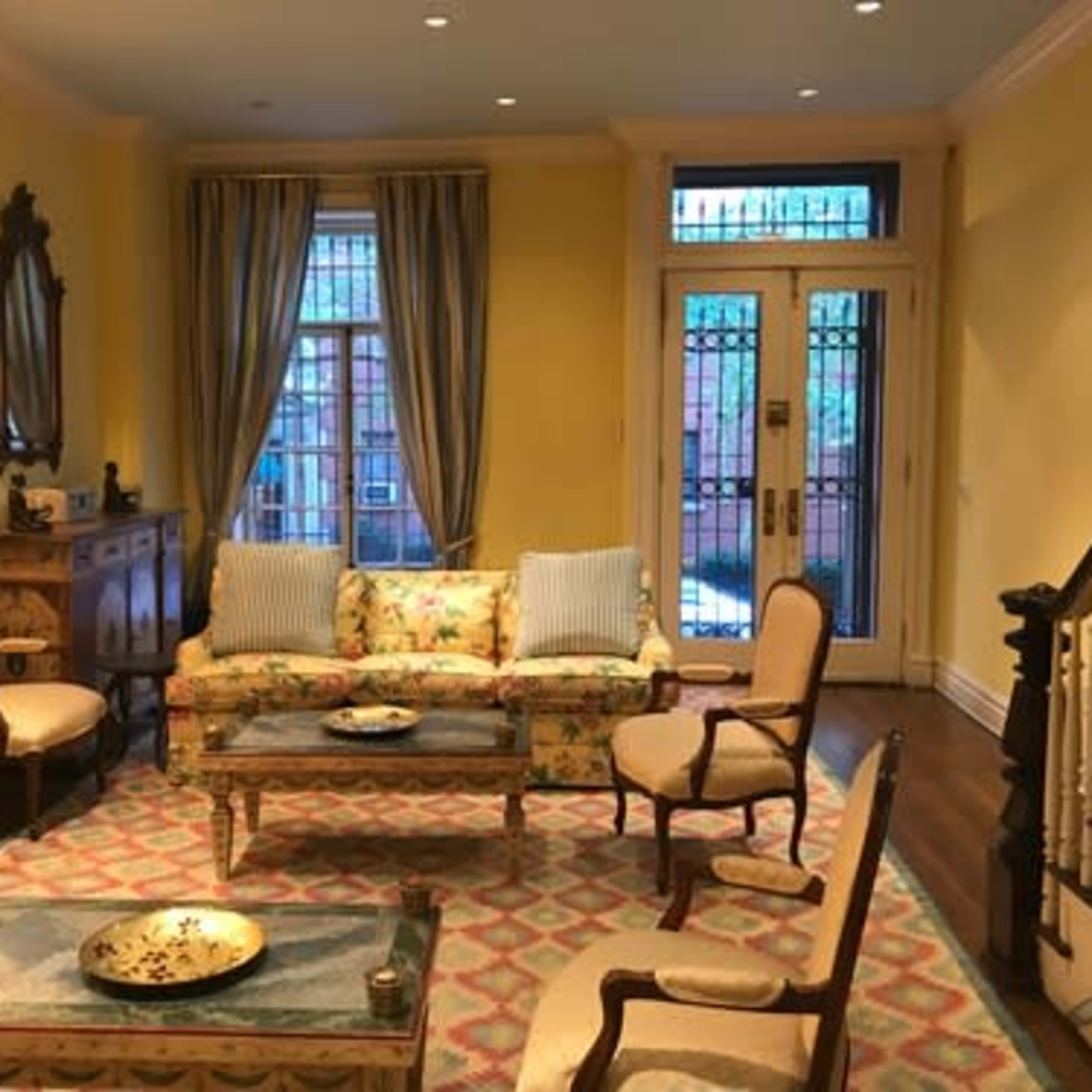 The room features a seating area with floral-patterned furniture, a coffee table, and a decorative rug, alongside a staircase and large windows.