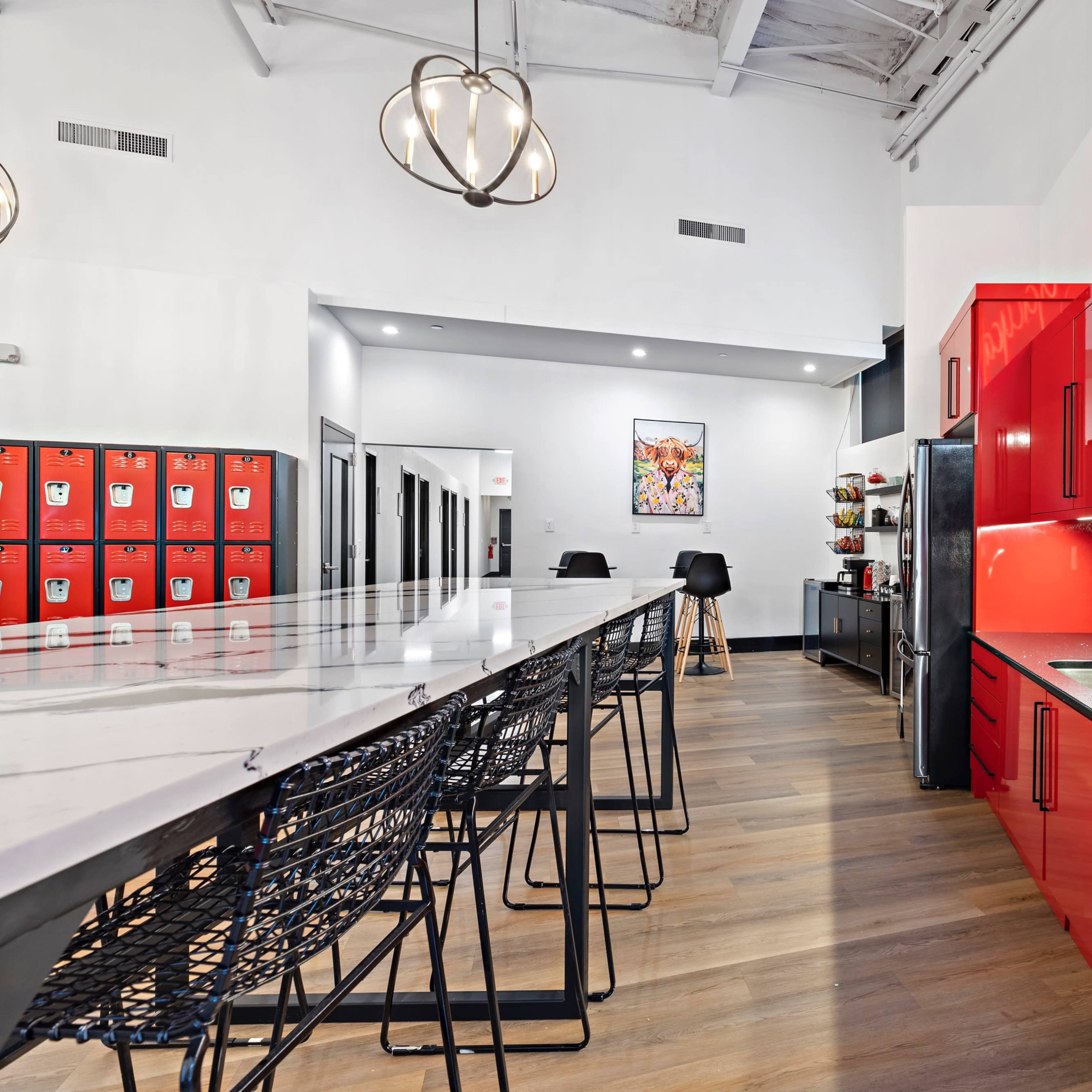 A modern kitchen with red cabinets and a long countertop, featuring bar stools, a refrigerator, and decorative storage lockers visible in the background.