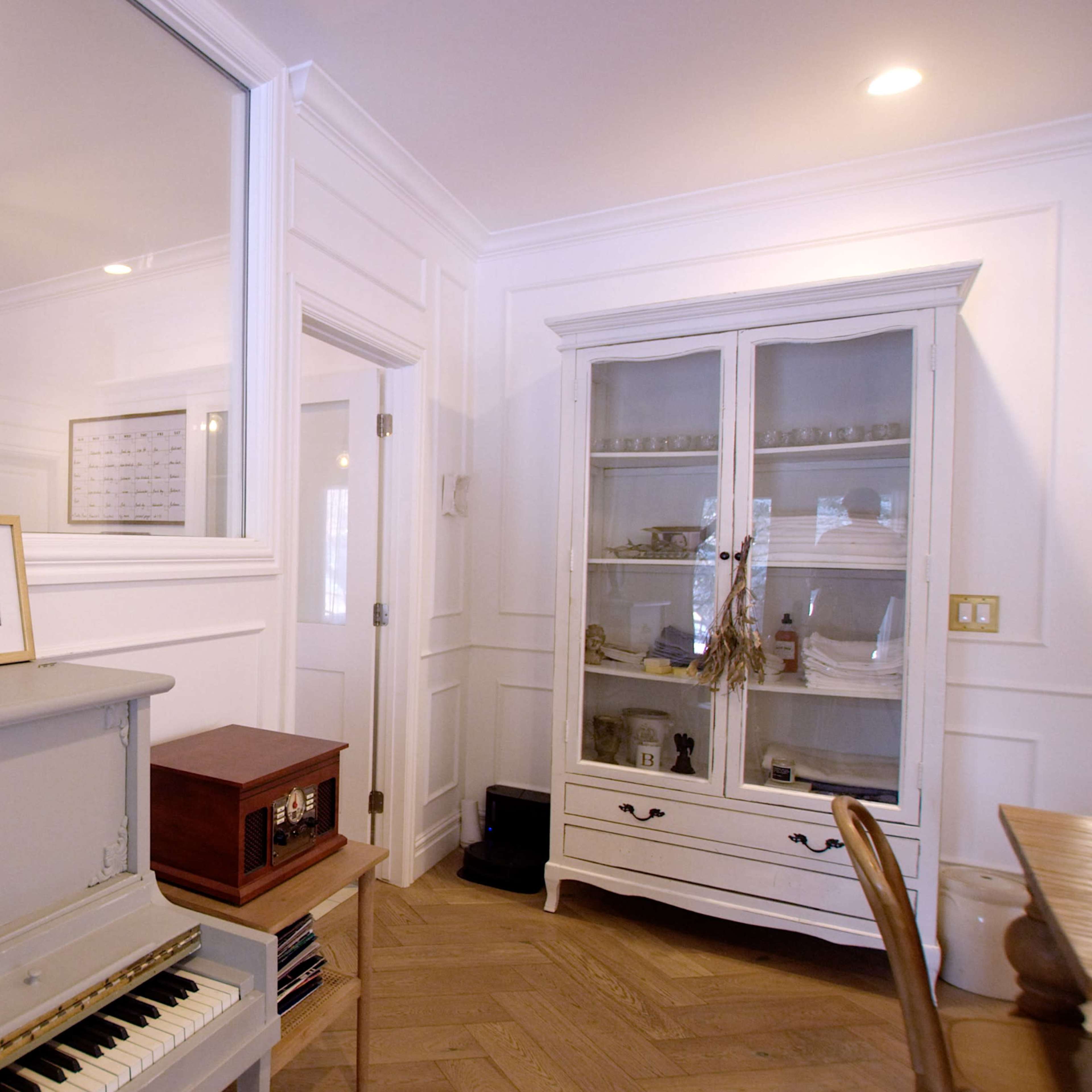 A room with a gray piano, a white cabinet displaying items, and a wall with white paneling and a mirror.