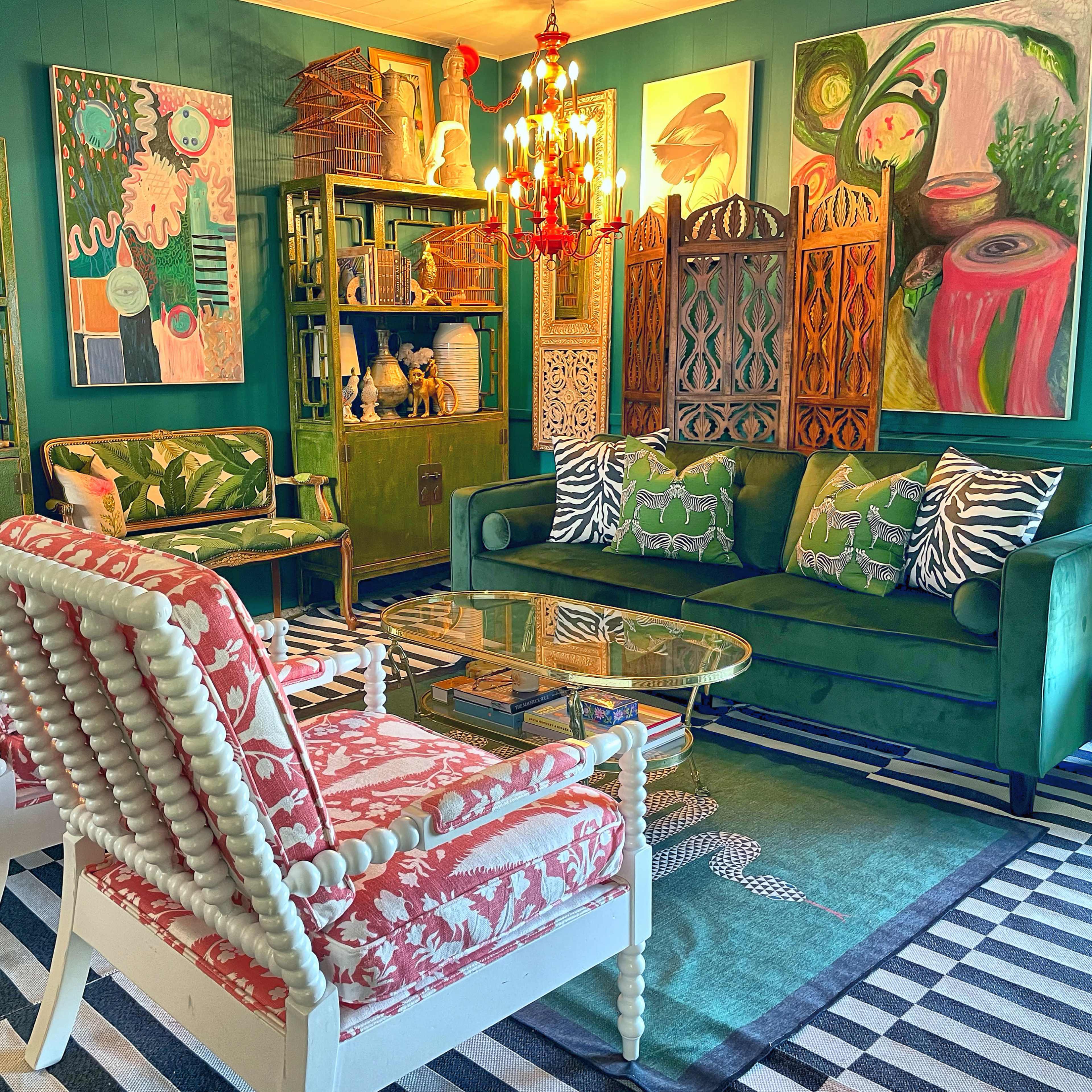 A colorful living room with green velvet sofas, patterned chairs, a glass coffee table, and various artworks displayed on the walls.