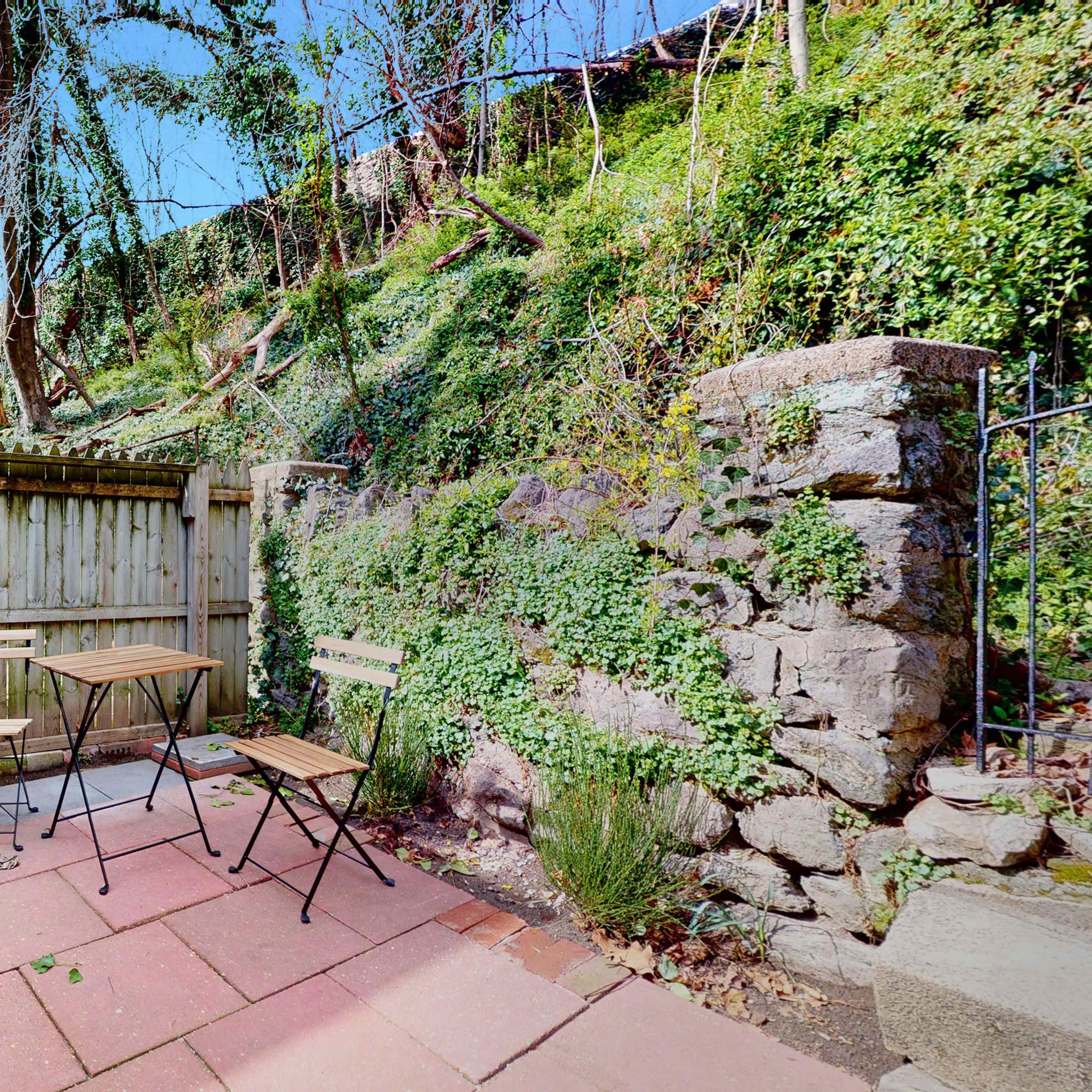 A small patio area features a table and two chairs against a backdrop of a wooden fence and a sloped, green hillside.