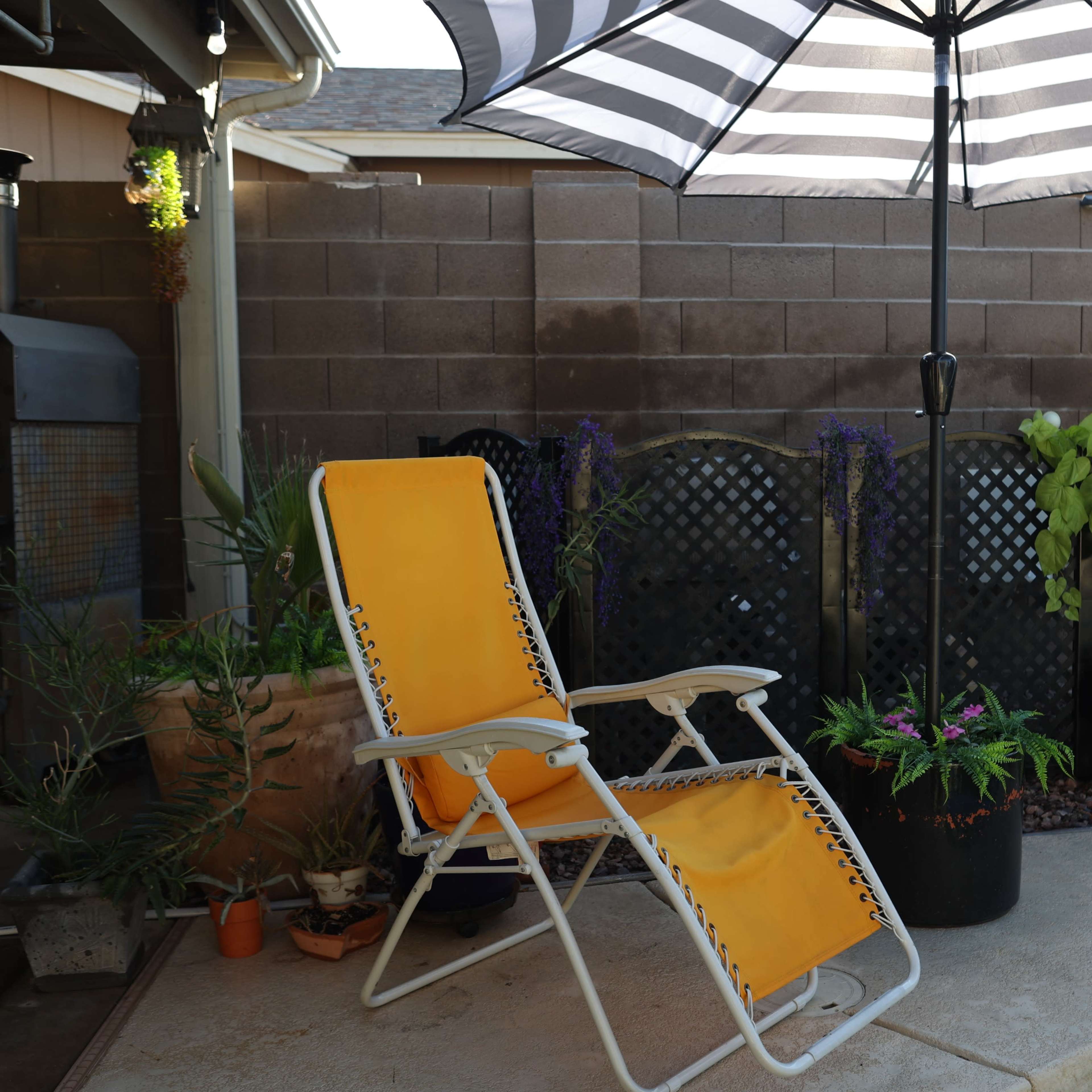A yellow lounge chair sits under a striped umbrella beside a pool, with potted plants and a brick wall in the background.