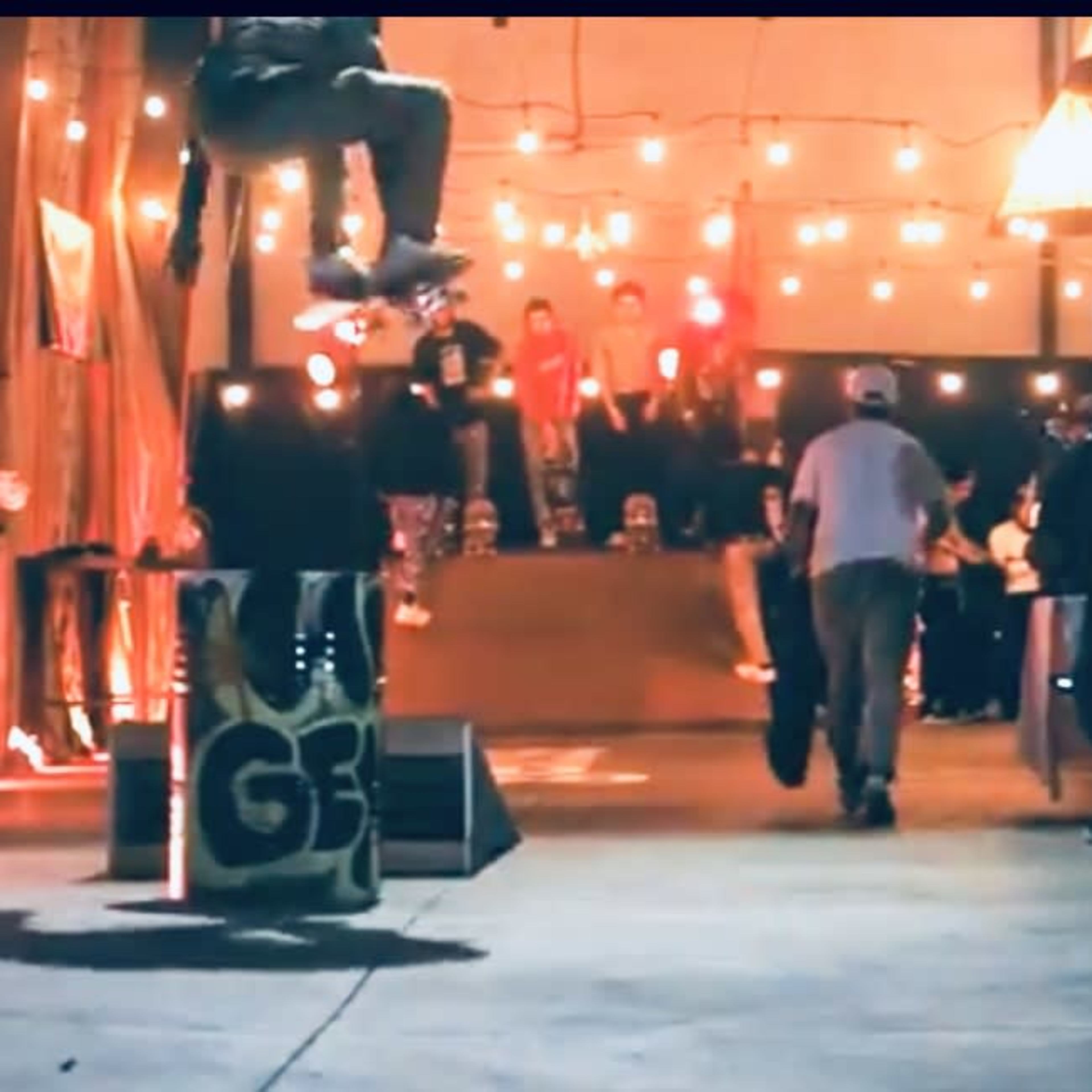 A skateboarder performs an aerial trick above a trash can in a dimly lit indoor skatepark filled with spectators.