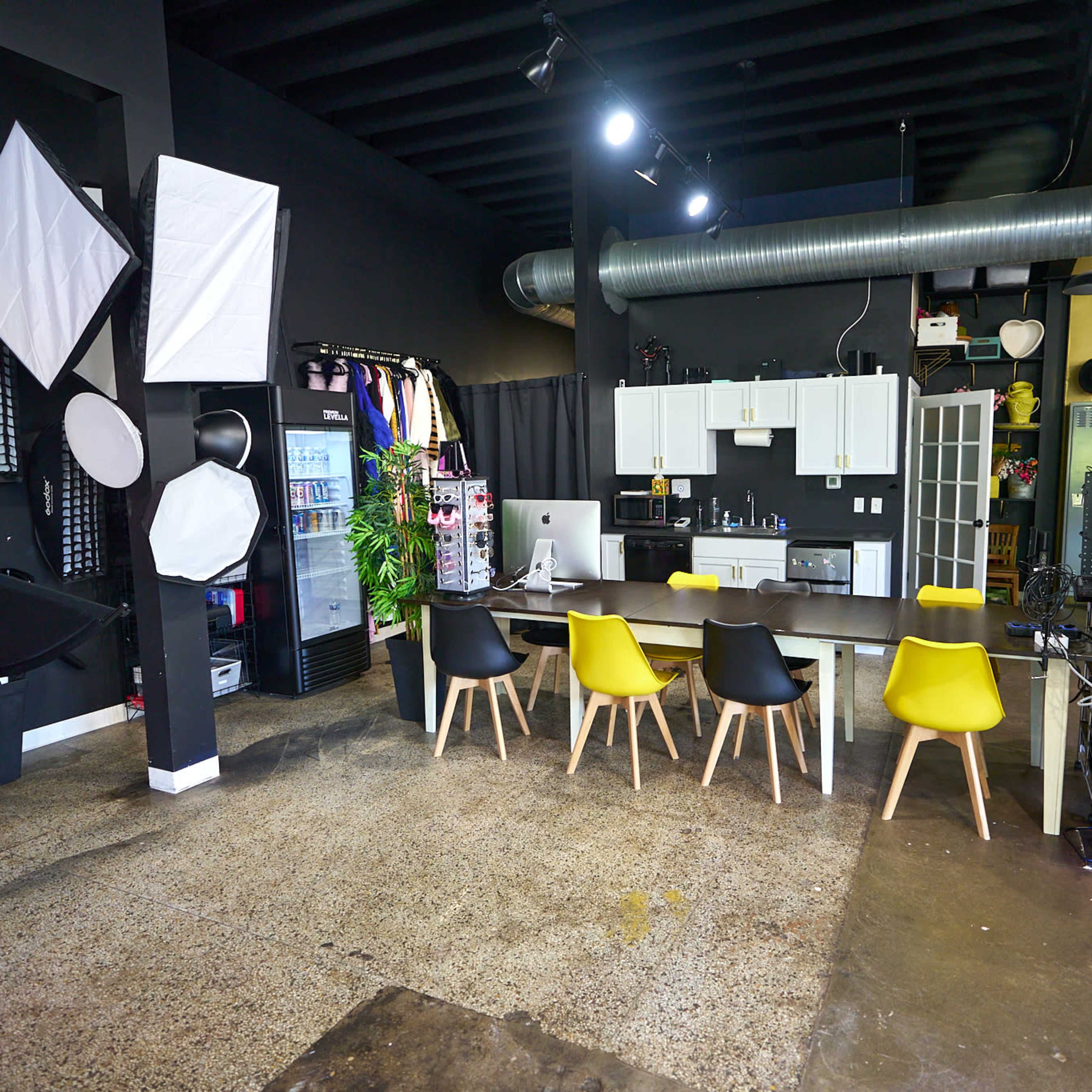 A modern workspace features a long table with colorful chairs, a kitchenette, and photography equipment along the walls in a black-painted room.