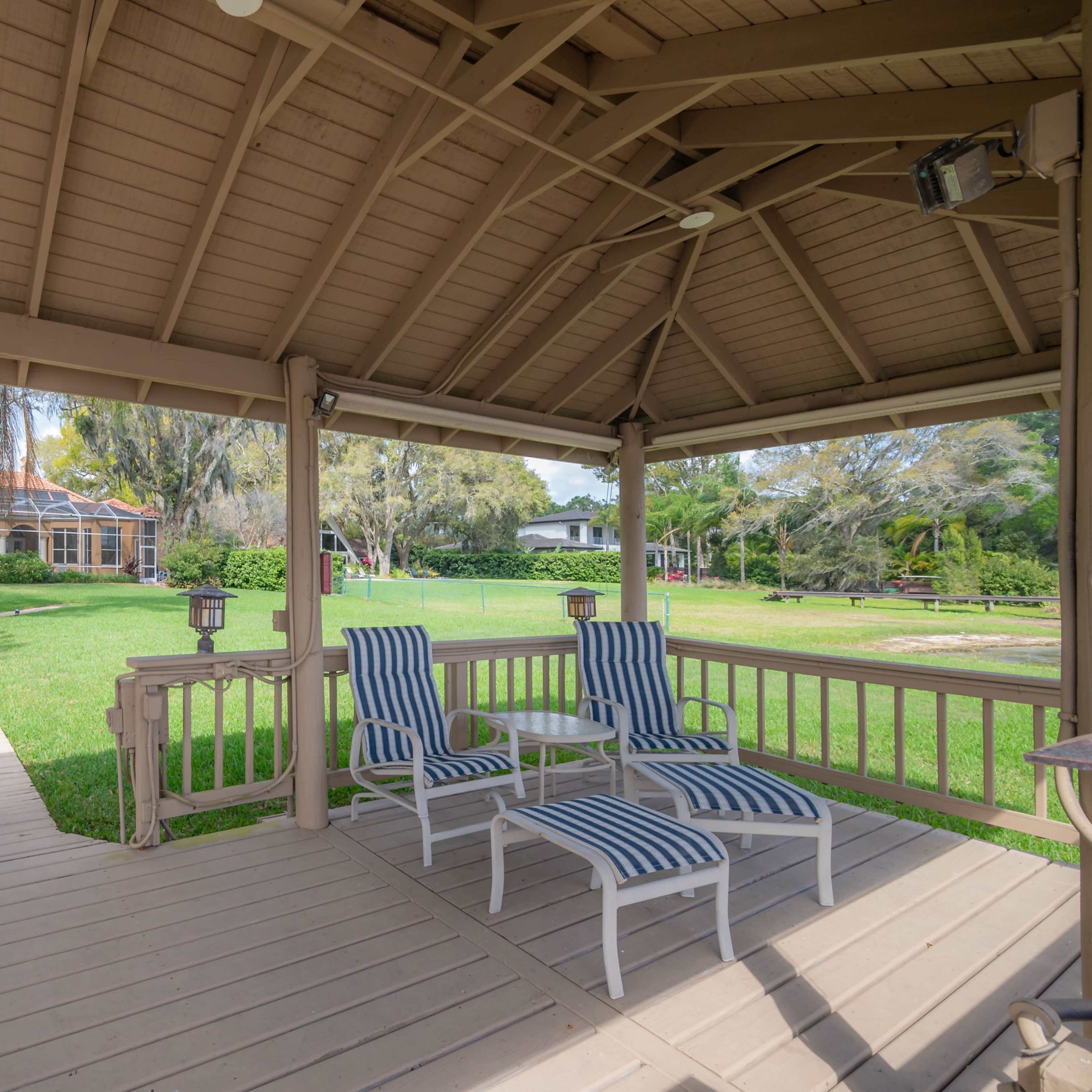 A covered deck features two striped lounge chairs and a small table, overlooking a grassy area and a path leading to a house in the background.