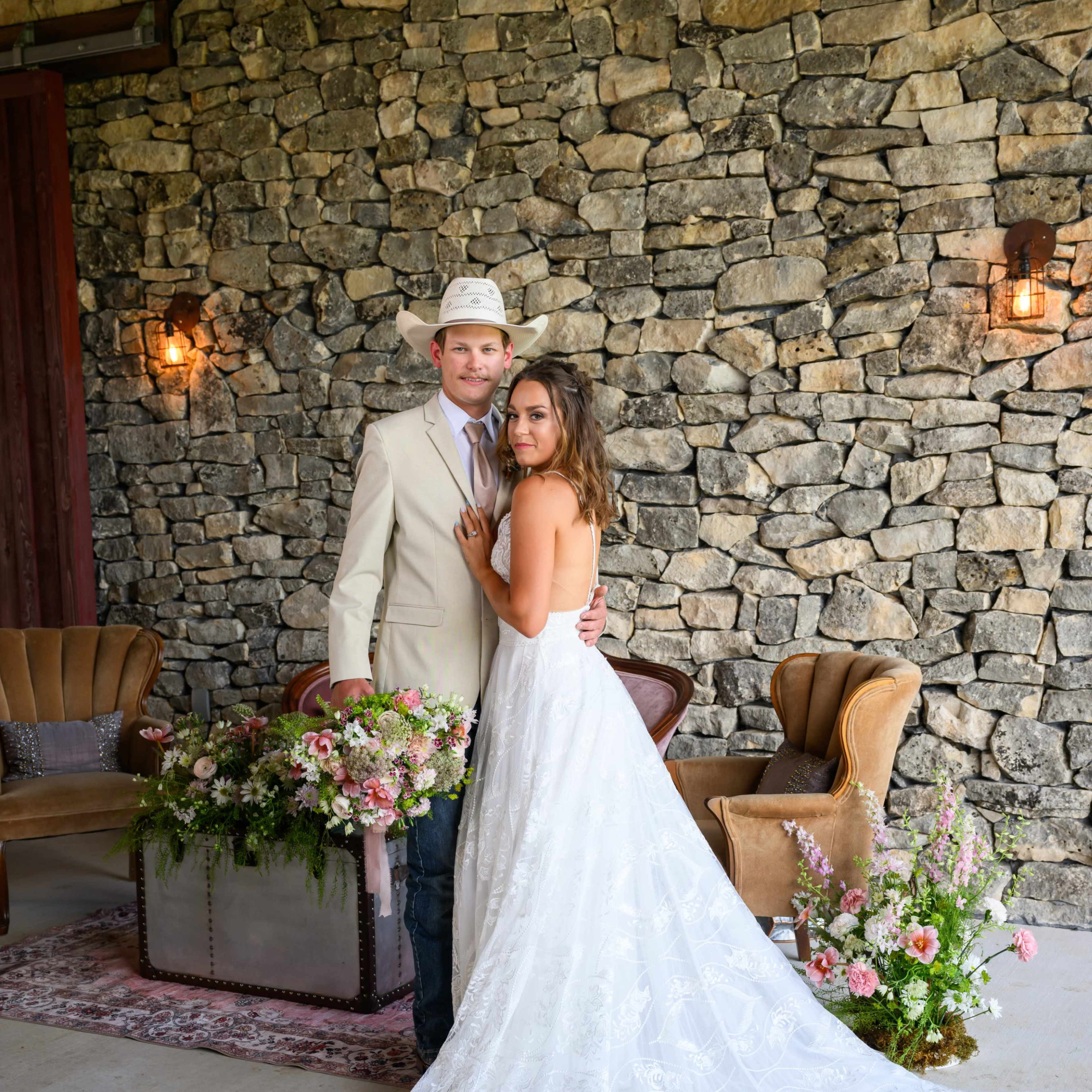 A couple poses together in front of a stone wall, with the woman in a wedding dress and the man in a light suit and hat, surrounded by floral arrangements and vintage furniture.