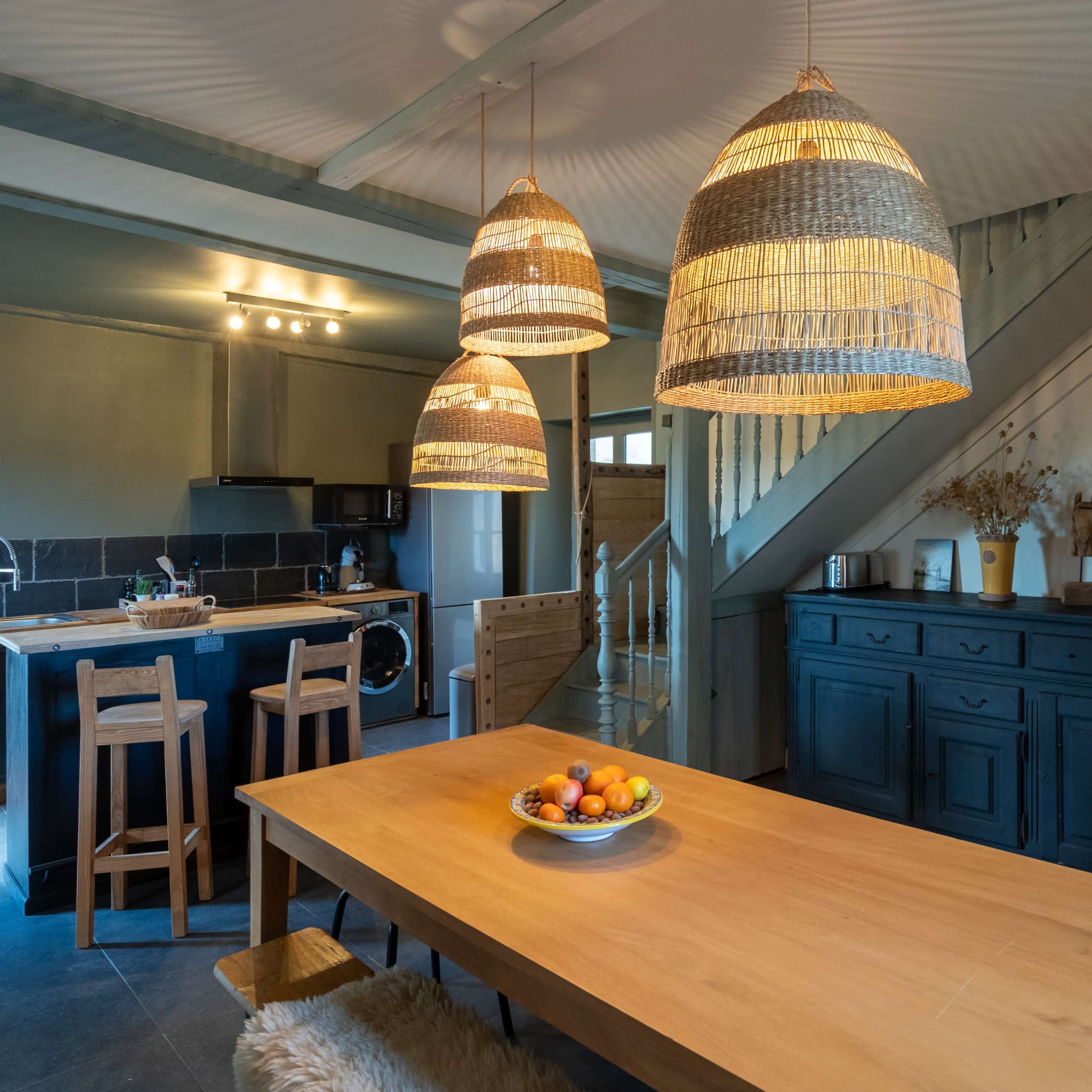 A spacious kitchen features a wooden dining table with a fruit bowl, three pendant lights, and a staircase leading to an upper level.