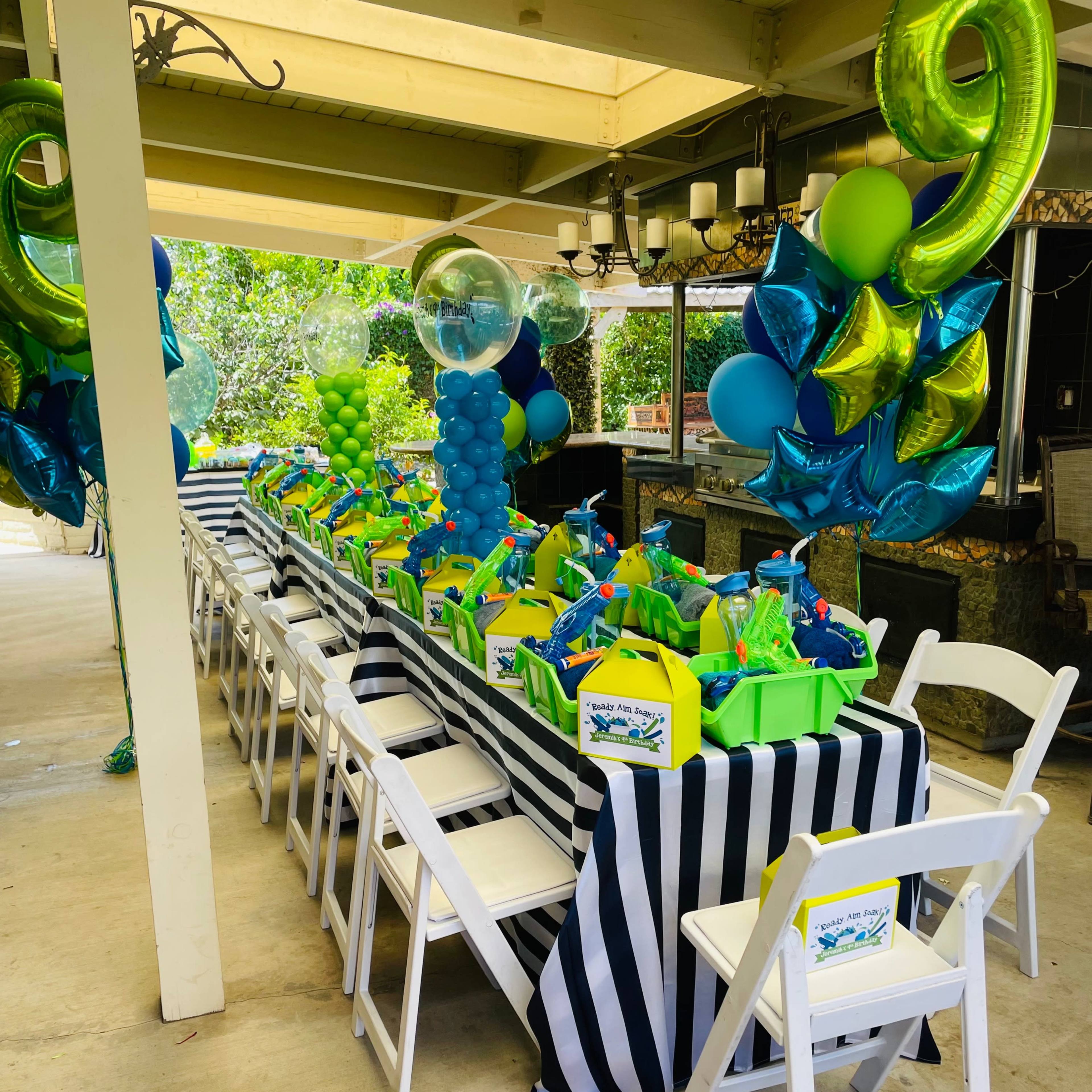 A long table is decorated with green and blue party favors, balloons, and striped tablecloths in a covered outdoor space.