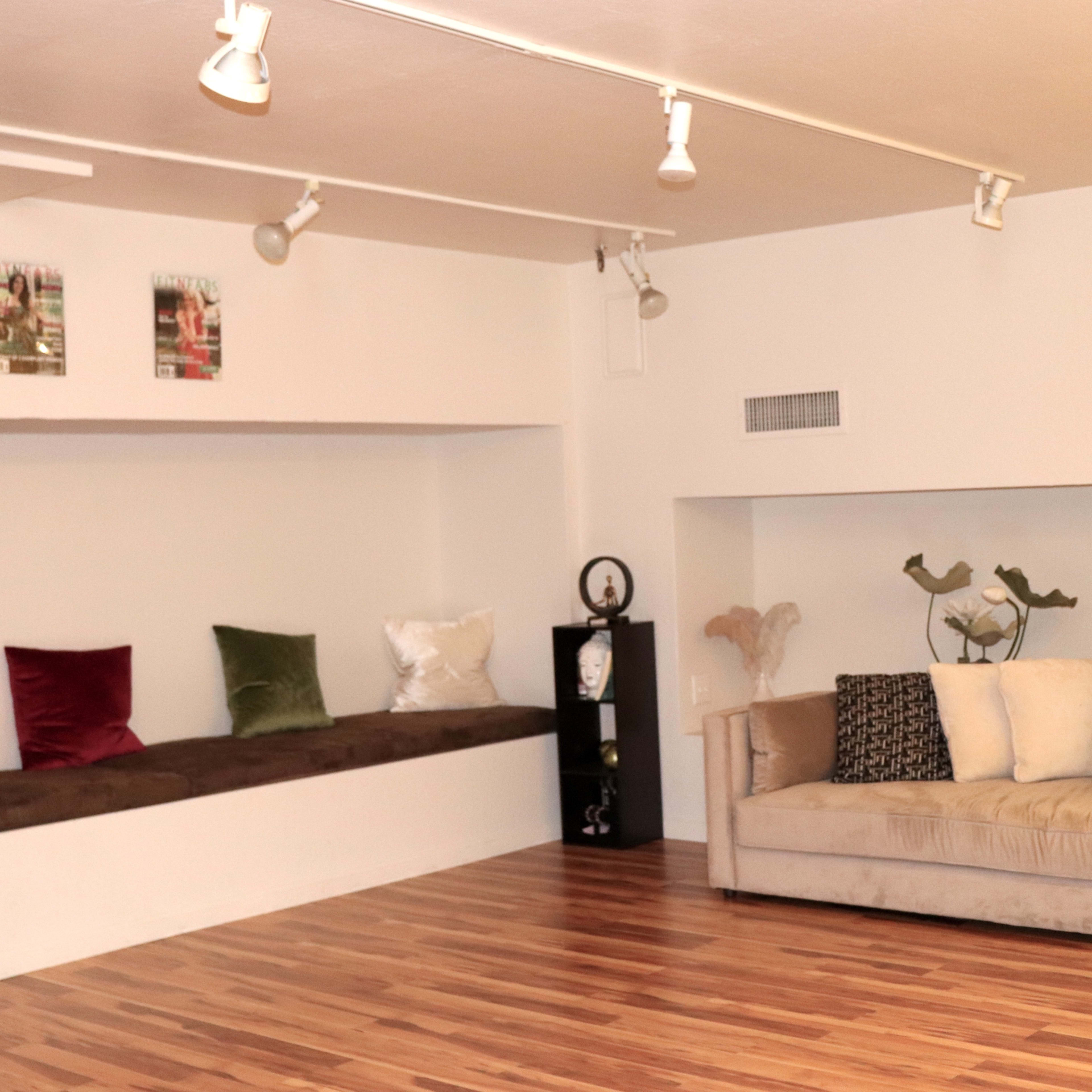 A spacious room with wooden flooring, featuring a long seating area with colorful cushions and a beige couch against the wall.