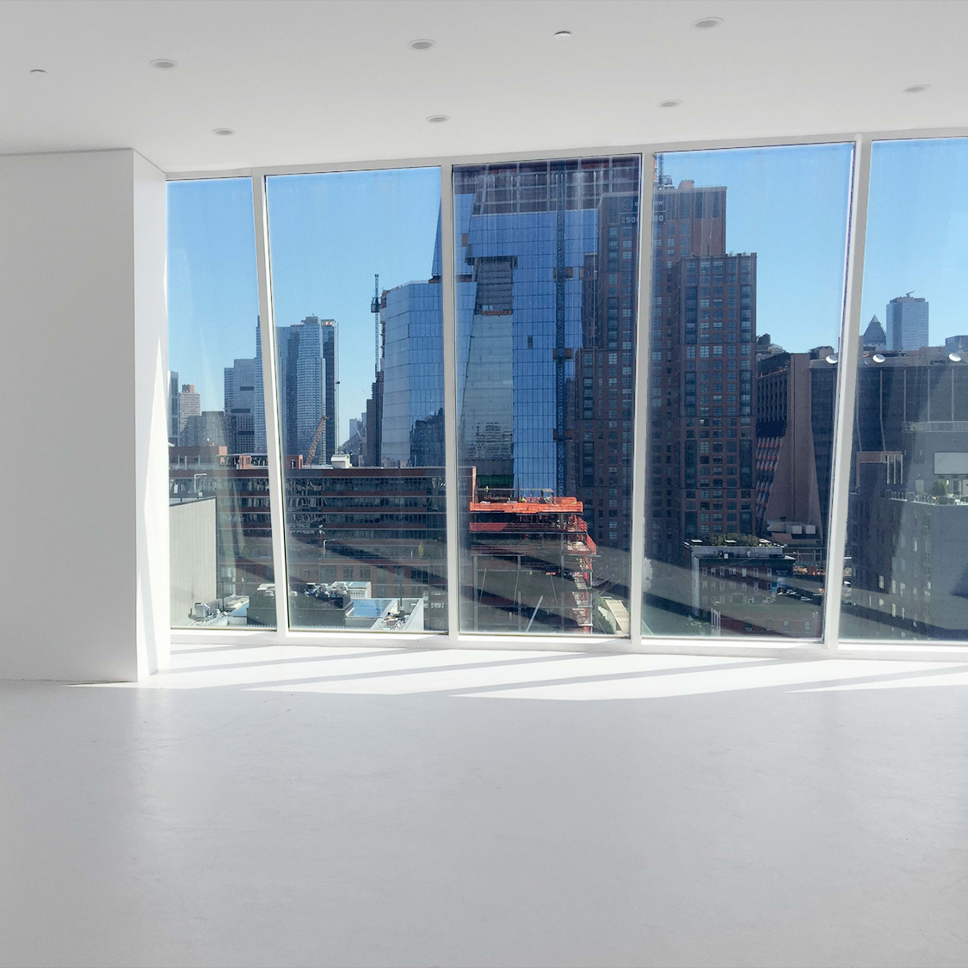 A spacious, minimalist room features large windows overlooking a city skyline with tall buildings and construction sites.