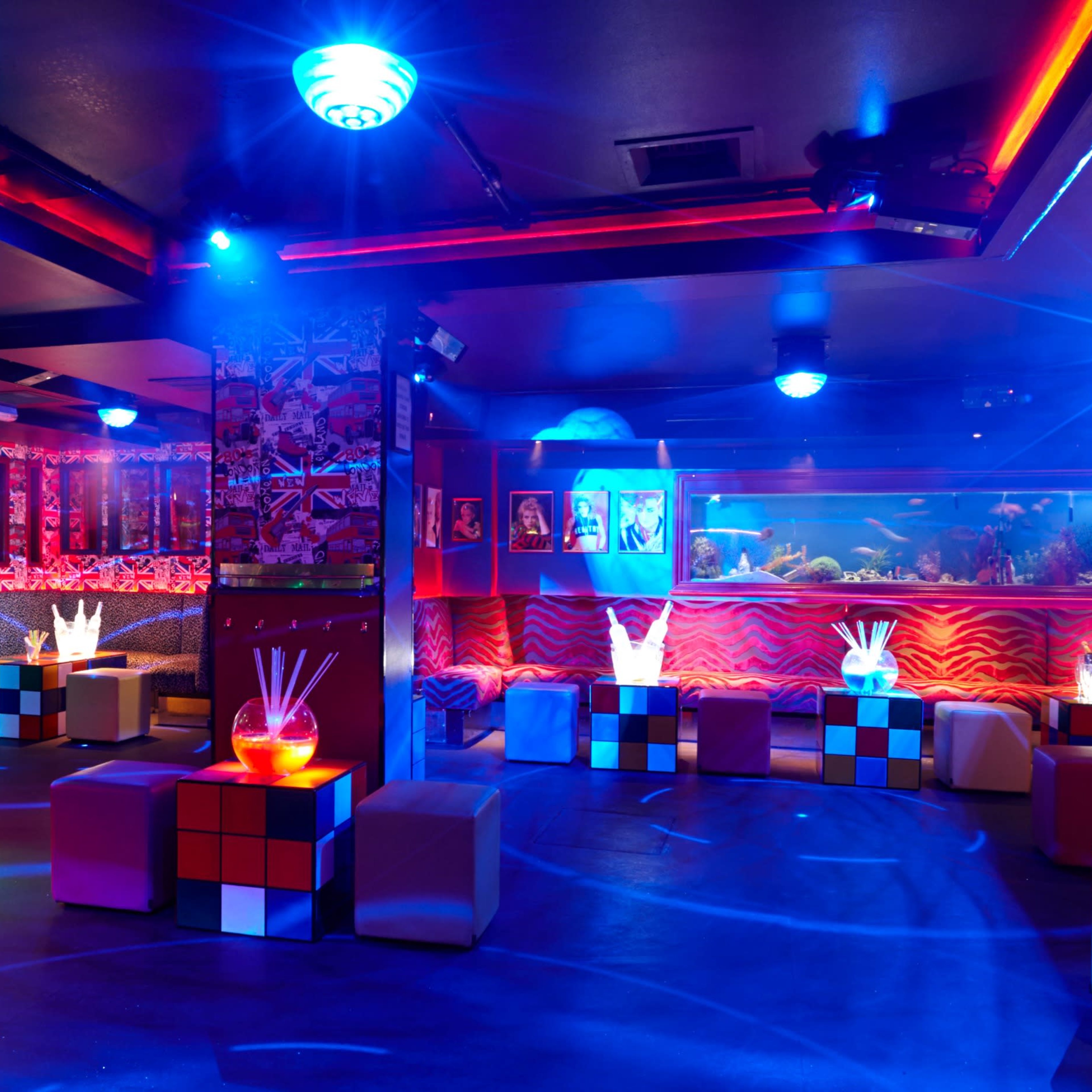 A vibrant nightclub features colorful lighting, a bar with illuminated drinks, and modern seating arranged around cube-shaped tables.