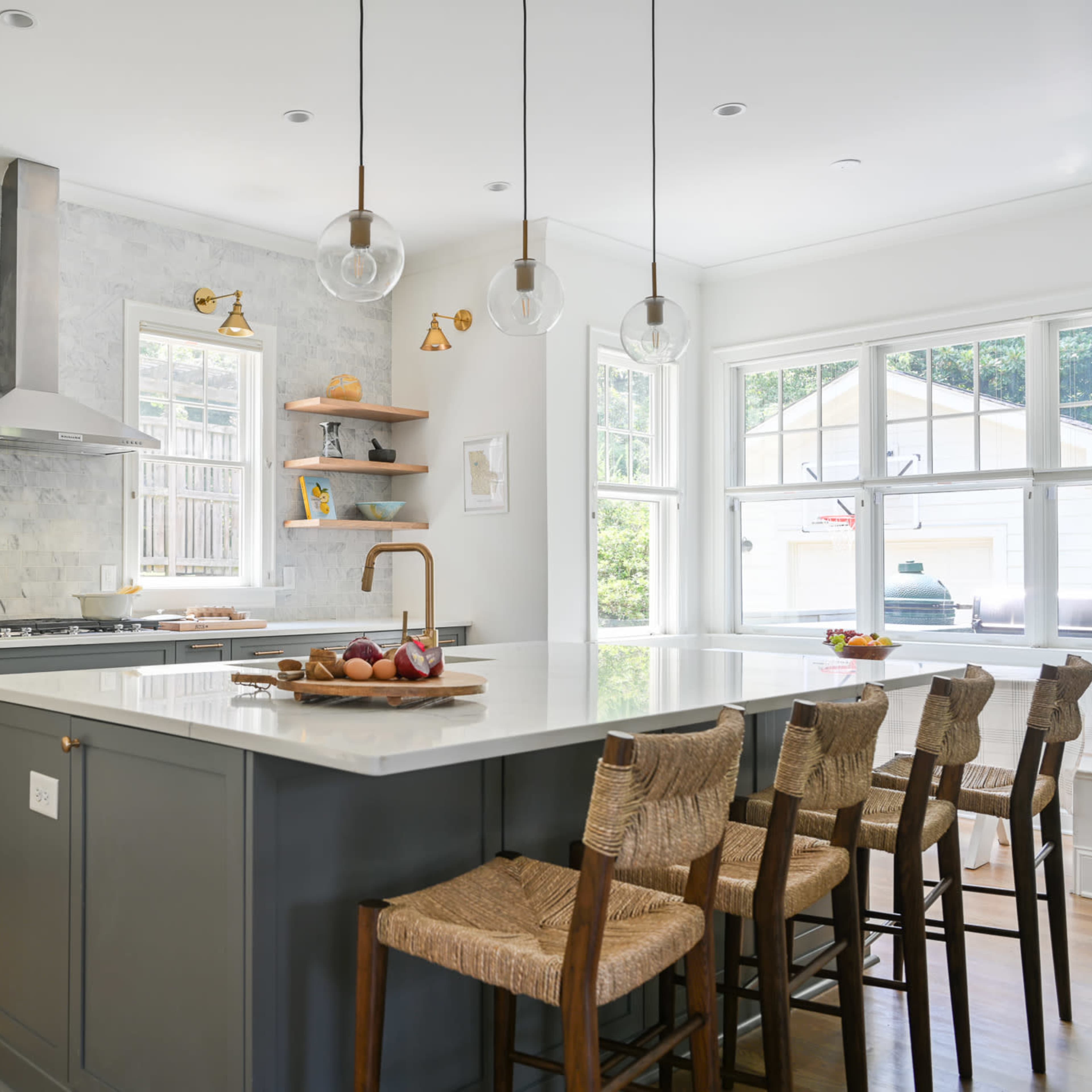 A modern kitchen features a large island with eight woven bar stools, marble countertops, stainless steel appliances, and a backsplash of light-colored tiles.