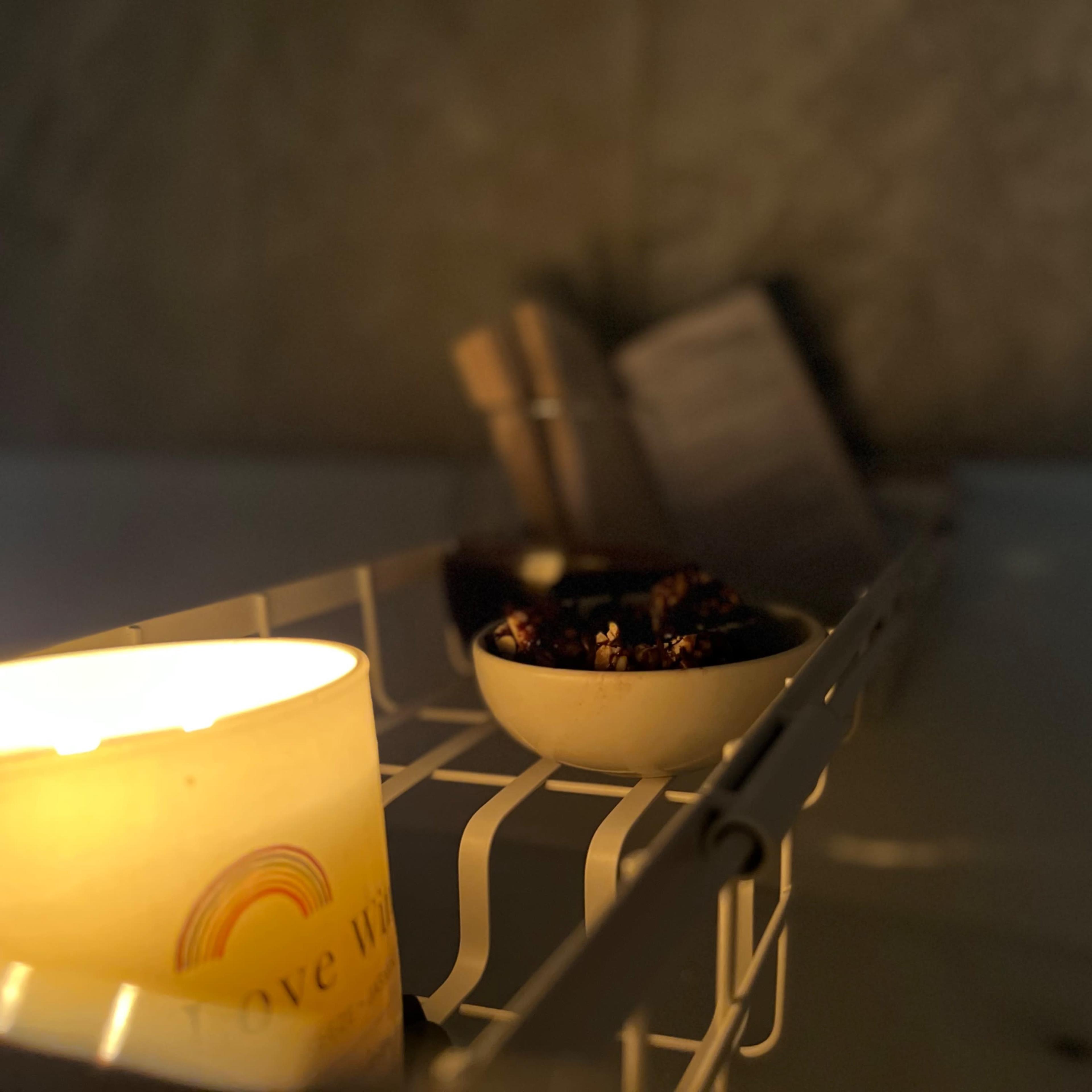A lit candle and a small bowl of dark food items are set on a white rack near a bathtub.