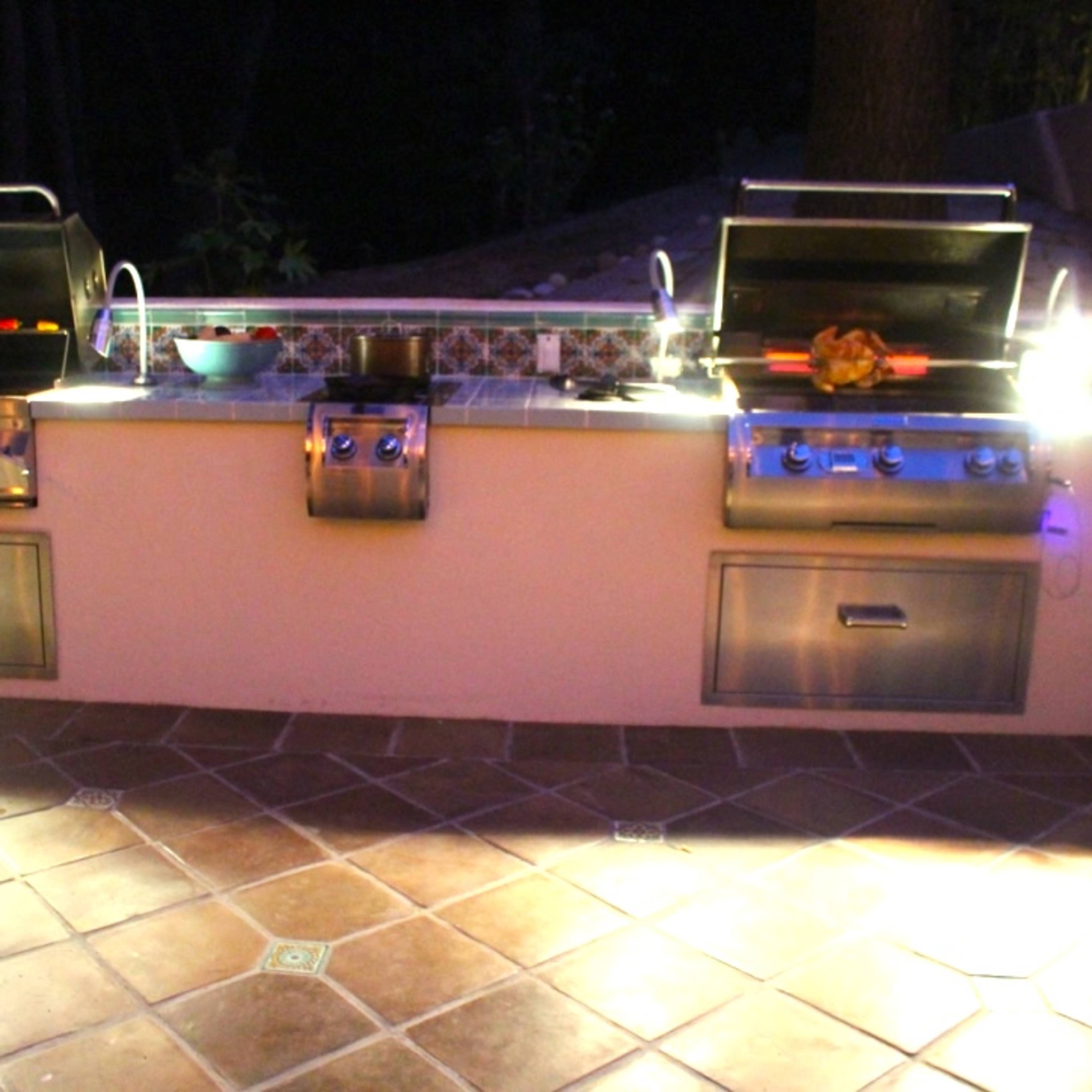 A well-equipped outdoor kitchen featuring two stainless steel grills and a small sink, illuminated by ambient lighting against a tiled backdrop.