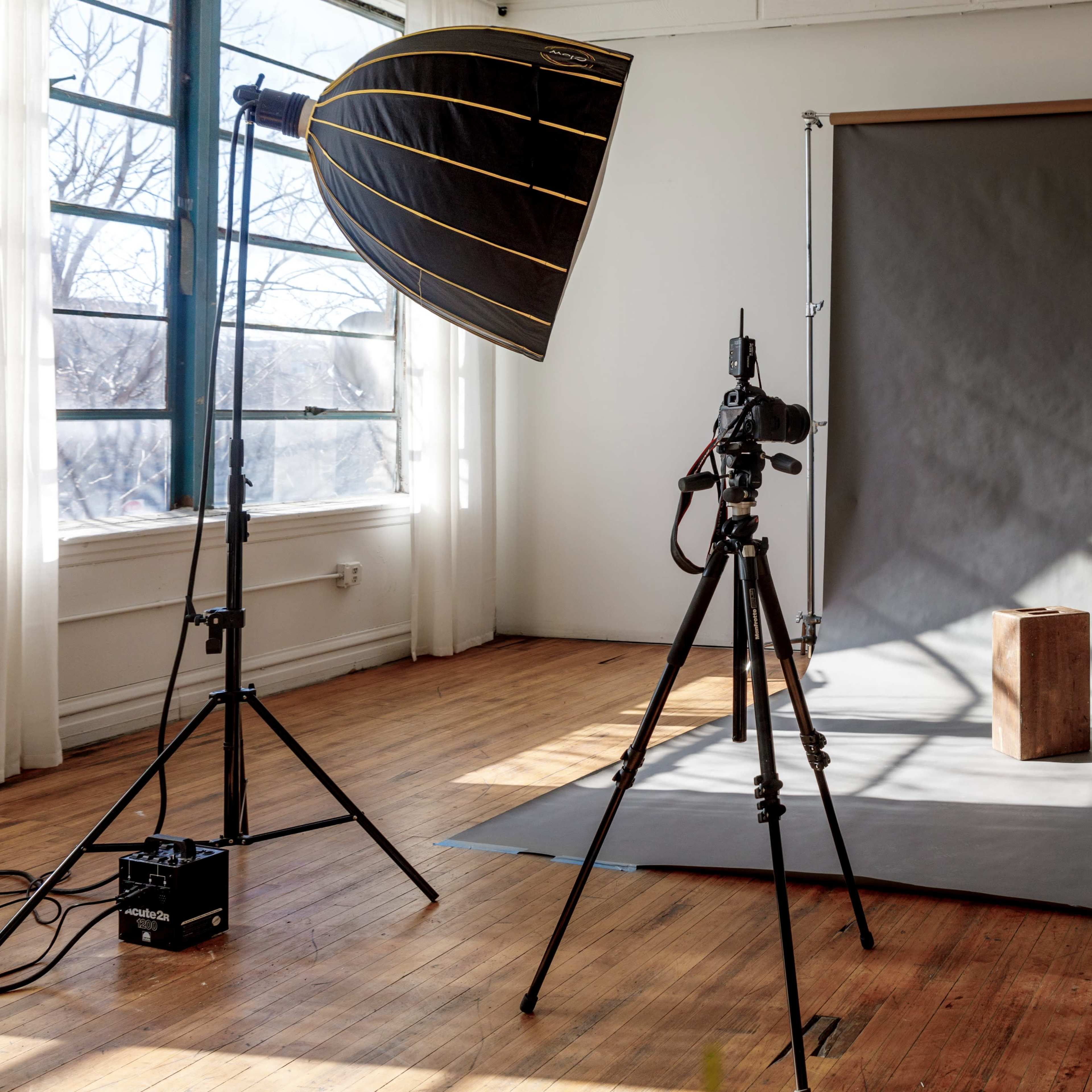 A photography studio with a wooden backdrop, a camera on a tripod, a large softbox light, and a reflector on either side.