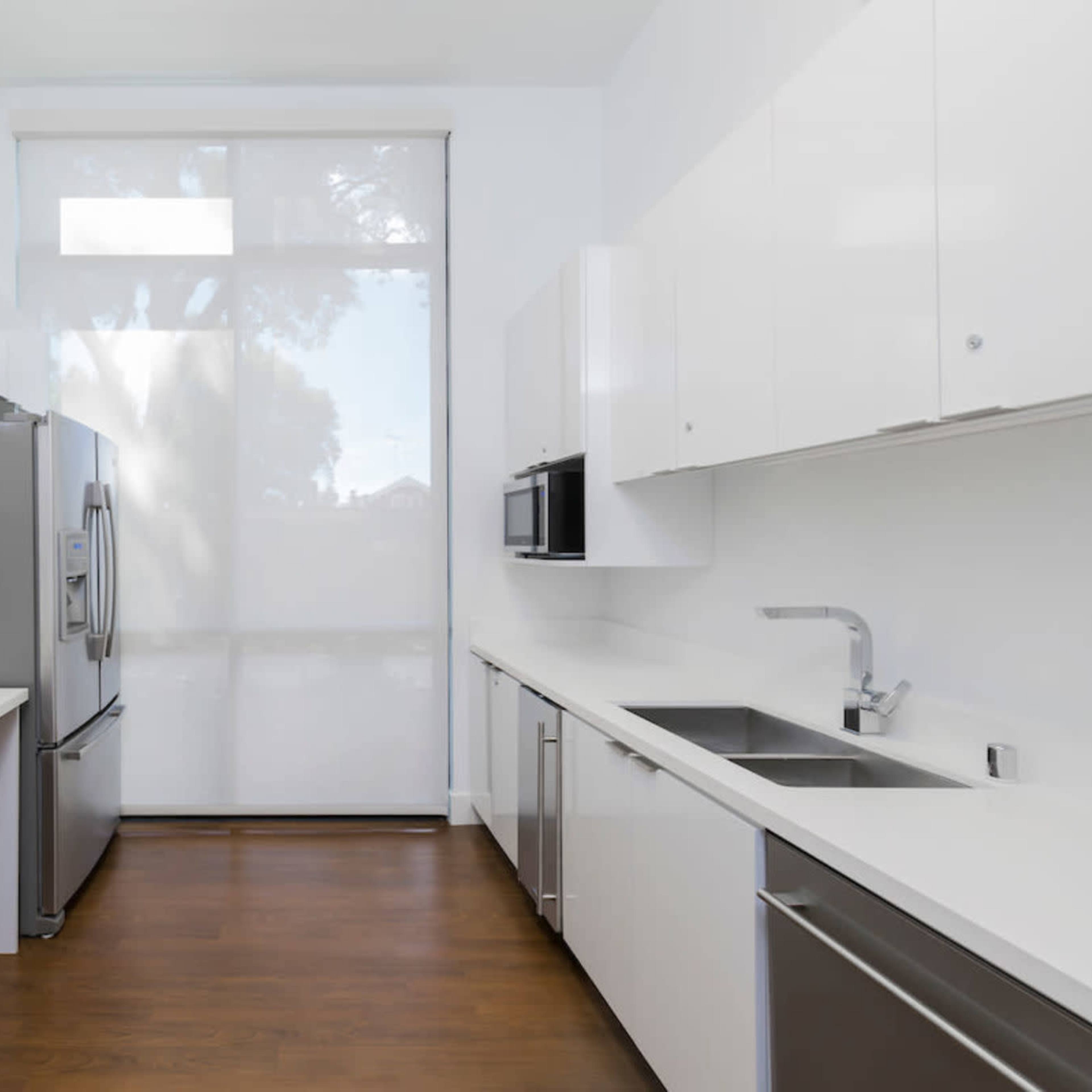 A modern kitchen features sleek white cabinetry, stainless steel appliances, and a large window allowing natural light to enter the space.