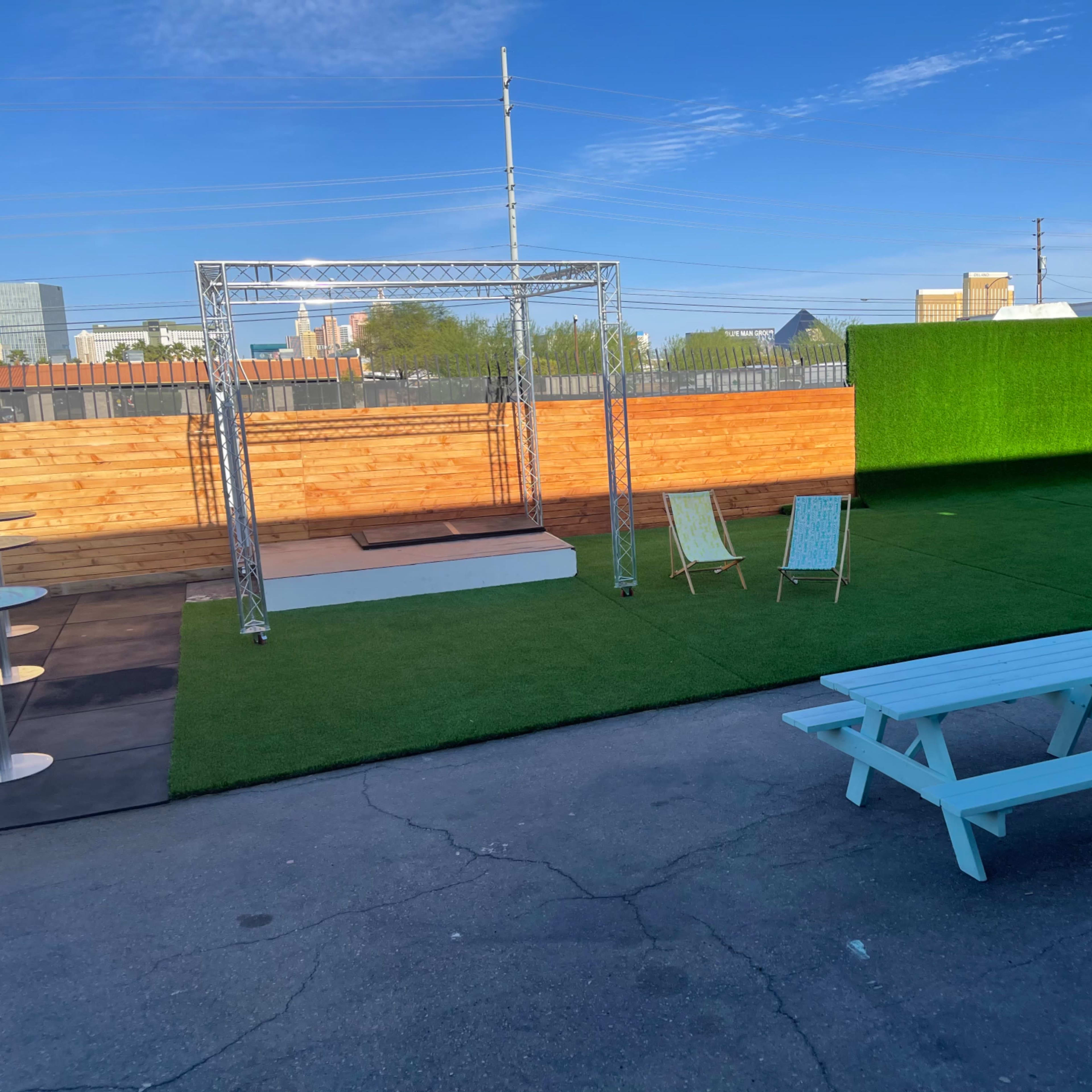 An outdoor venue featuring a grassy area with two light blue picnic tables, two chairs, and a performance stage framed by a green backdrop.